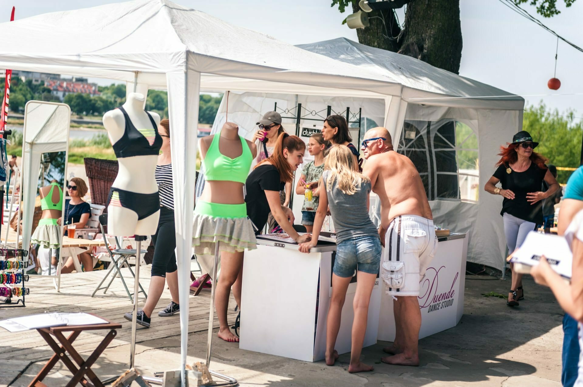 People gather at a stand under a white canopy at an outdoor market. On display are mannequins dressed in swimwear and summer clothes. The scene, part of a photo essay of the fair, includes several people browsing and chatting, while trees and other booths are visible in the background.  