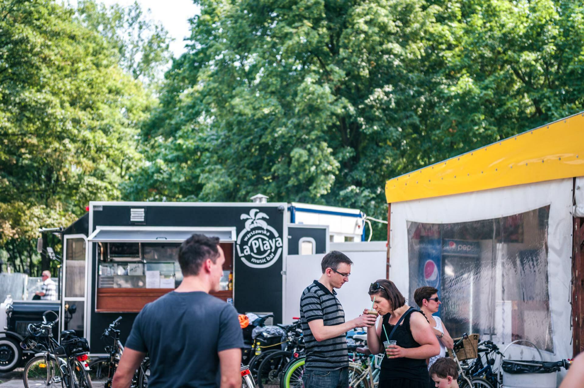 Photo report from the fair: An outdoor scene with several people in casual attire gathered around food trucks and a tent. One man is serving a drink to a woman, a child is standing nearby, and bicycles and trees can be seen in the background, indicating a relaxed, social atmosphere. A sign on one of the food trucks reads "La Playa.  