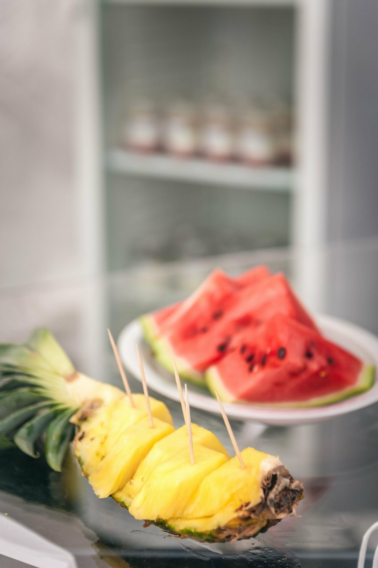 A plate of sliced watermelon and halved pineapple with skewers are set on a glass table, perfectly capturing the essence of the colorful spread. Shelves of small jars, presumably containing desserts or condiments, are slightly smudged in the background of this photo report from the fair. 