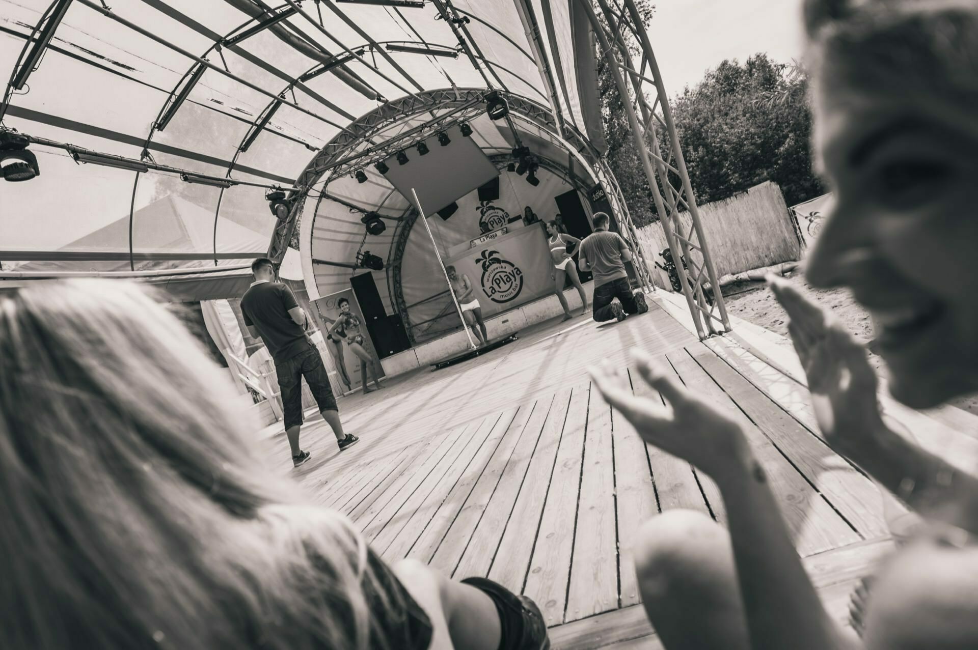 Black and white image of a small outdoor stage. Several people can be seen, some standing on the stage and some sitting in the foreground. The stage has a curved roof, backdrop and sound system with overhead lighting. The sunlit area captures every detail - a true photo-reportage of the fair.   