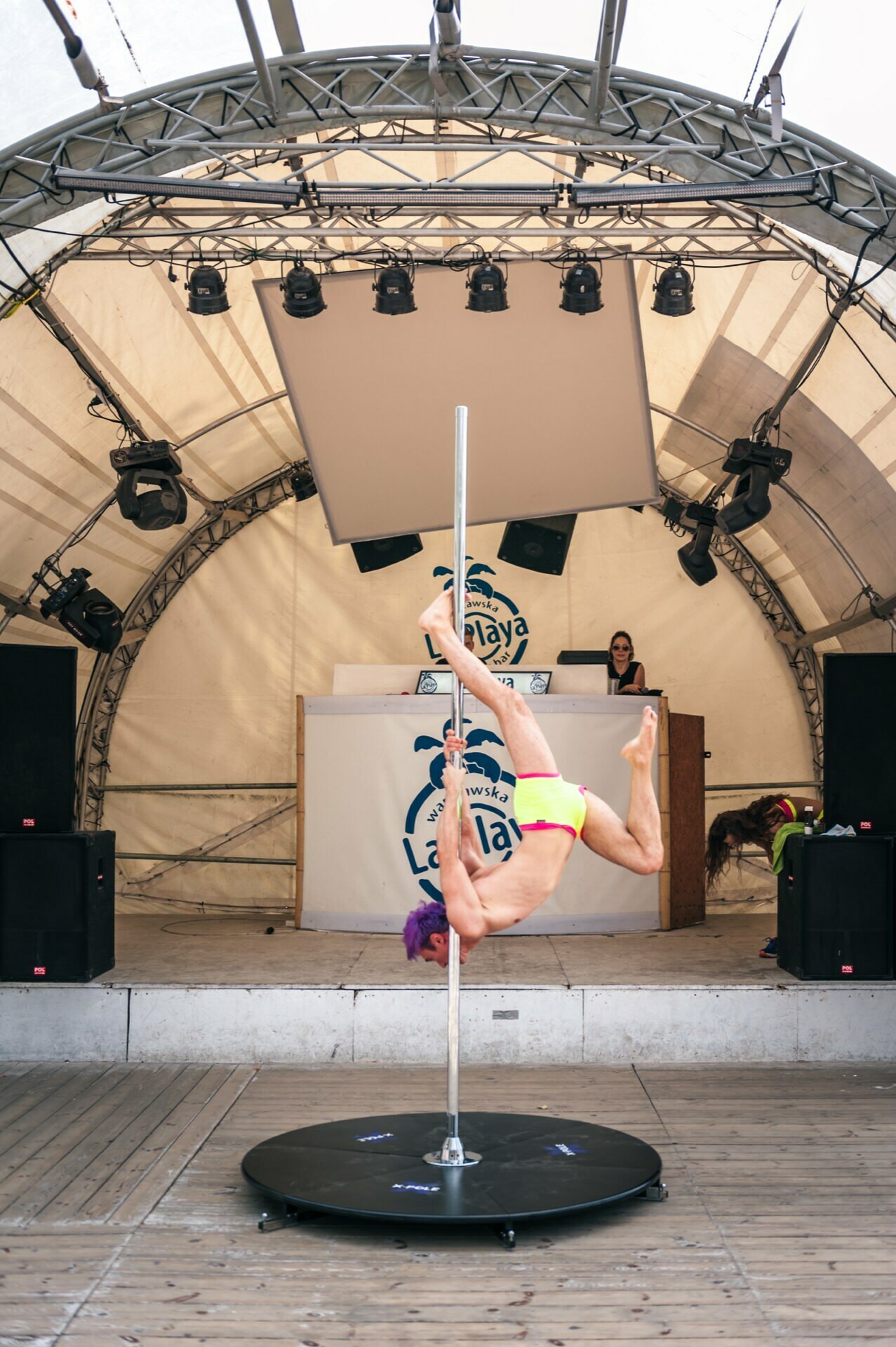 A person with colorful hair, dressed in neon shorts, performs an acrobatic pole dance on stage. A DJ stands at a booth in the background with speakers and a large screen behind them. The stage is set up inside a white tent with a metal frame, capturing the vivid essence of the photo fair.  