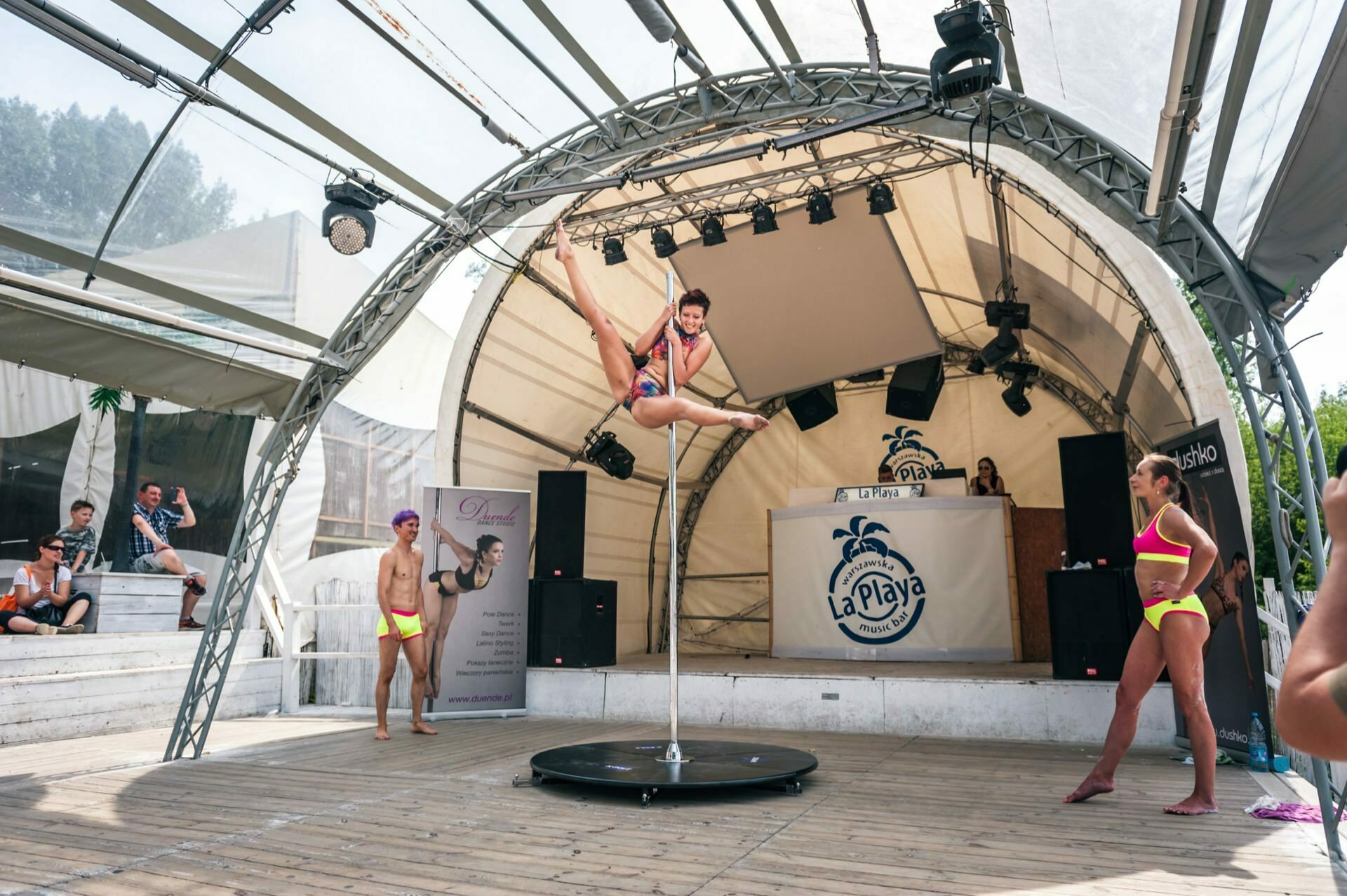 A person dressed in colorful athletic attire performs a pole dance on an outdoor stage under a canopy that reads "La Playa." Two other performers stand to the side in neon outfits, while a small audience watches the performance from a nearby staircase. The setup suggests a beach or summer event, perfect for a photo fair.  