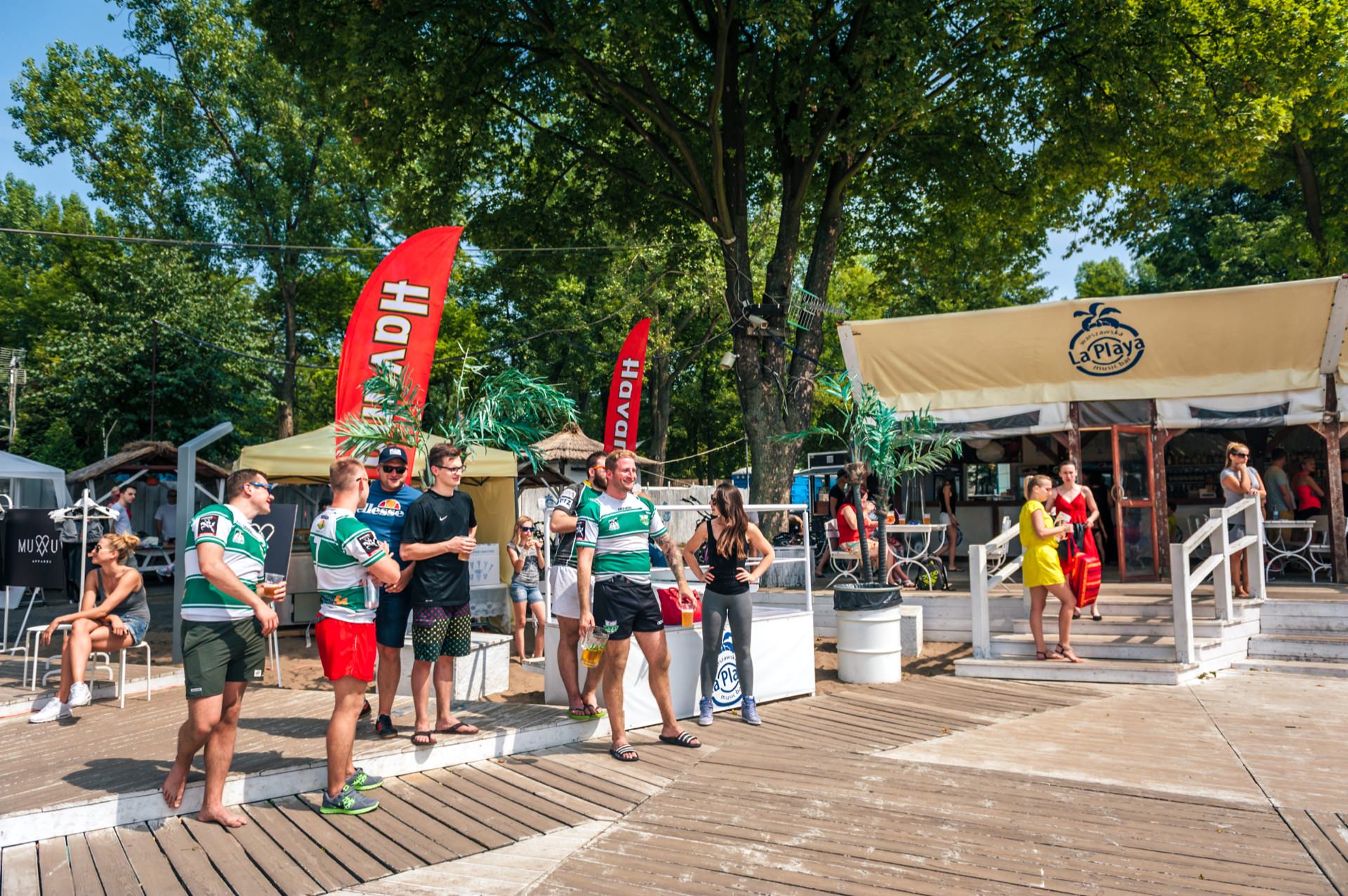 A group of people standing on a terrace with a beach bar and large red flags in the background. Some are wearing sportswear, others are wearing loose-fitting clothing. Trees and a building with a sign that reads "La Playa" are visible, providing the perfect backdrop for this photo essay of a fair in a beach- or lake-like location.  