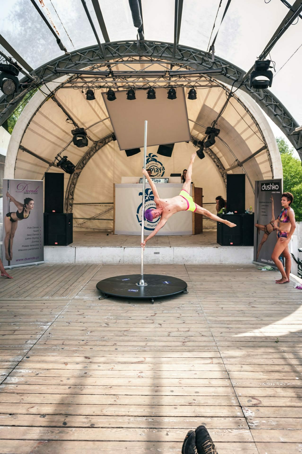 An artist demonstrates pole dance moves on an outdoor stage during a photo opportunity at the fair, assuming a horizontal position with one leg extended and the other lowered. The stage is equipped with speakers, a banner and posters, and a small audience watches the show. Trees can be seen in the background.  