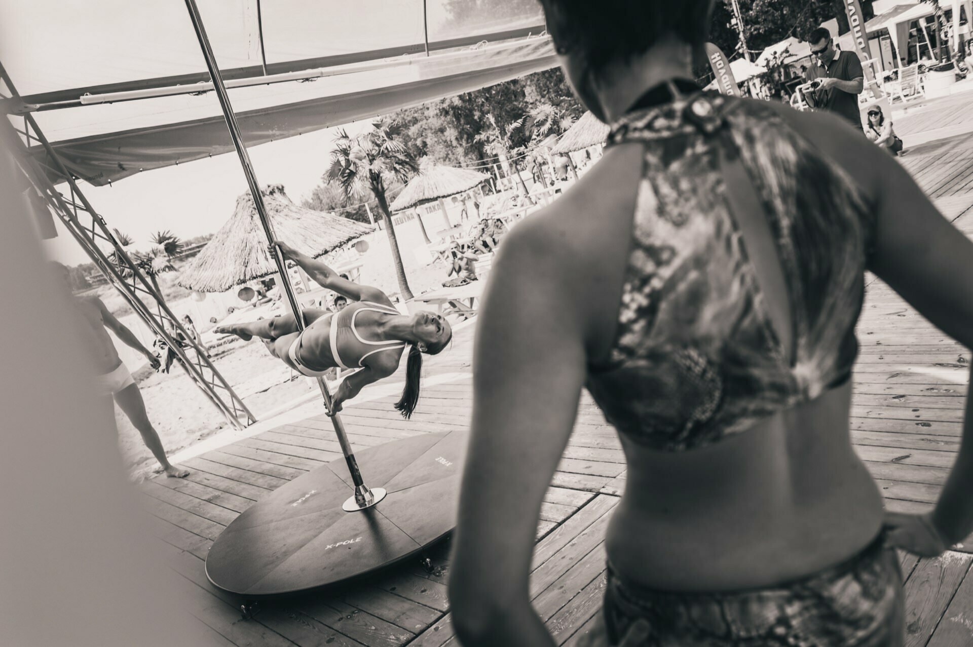 Black and white photo of a woman performing a pole dance on an outdoor stage while another woman looks with her back to the camera. The scene is set on a beach with straw umbrellas and people in the background. A captivating photo report from the fair capturing the vibrant atmosphere.  