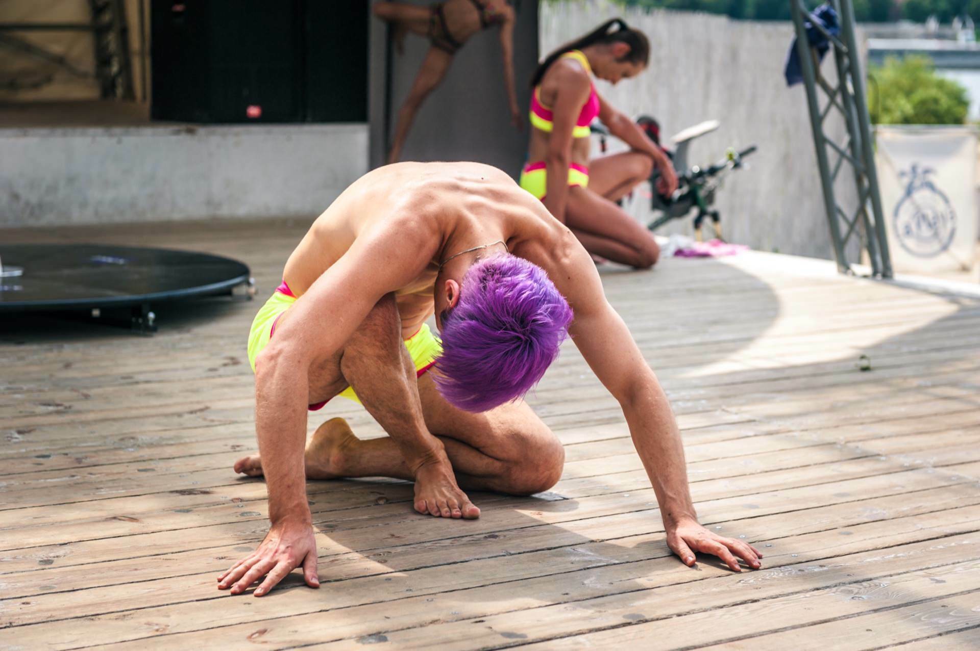 A person with purple hair, dressed in neon yellow and pink exercise clothes, is doing yoga or dancing on a wooden floor. Two other people in similar outfits can be seen in the background, one stretching and the other tweaking equipment - almost like an energizing photo-op at a trade show. 