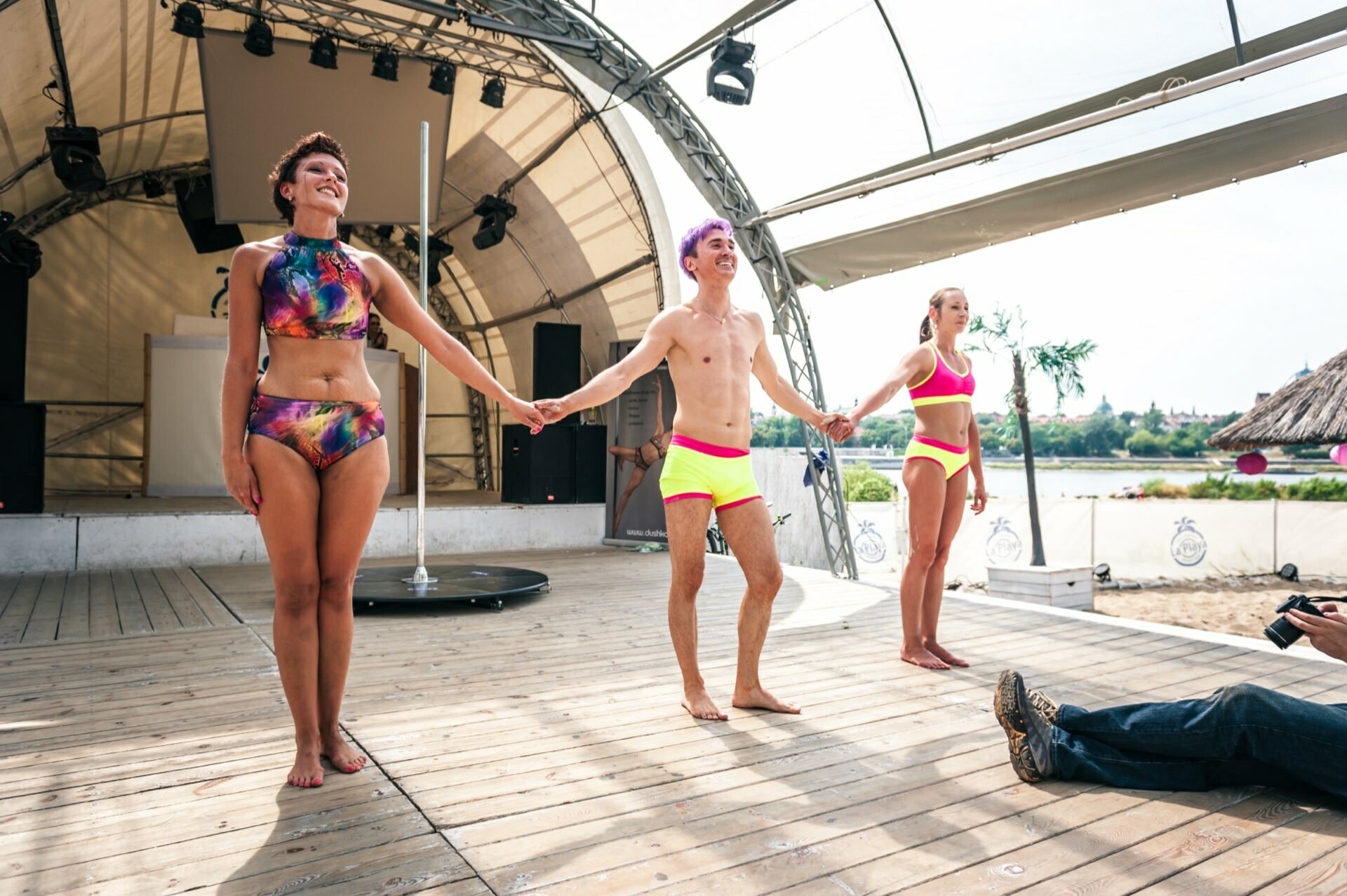 Three people in colorful bathing suits hold hands on a stage under a large canopy. The stage is a body of water with palm trees visible in the background. To the right, a photographer can be seen capturing this moment of photojournalism from the fair.  