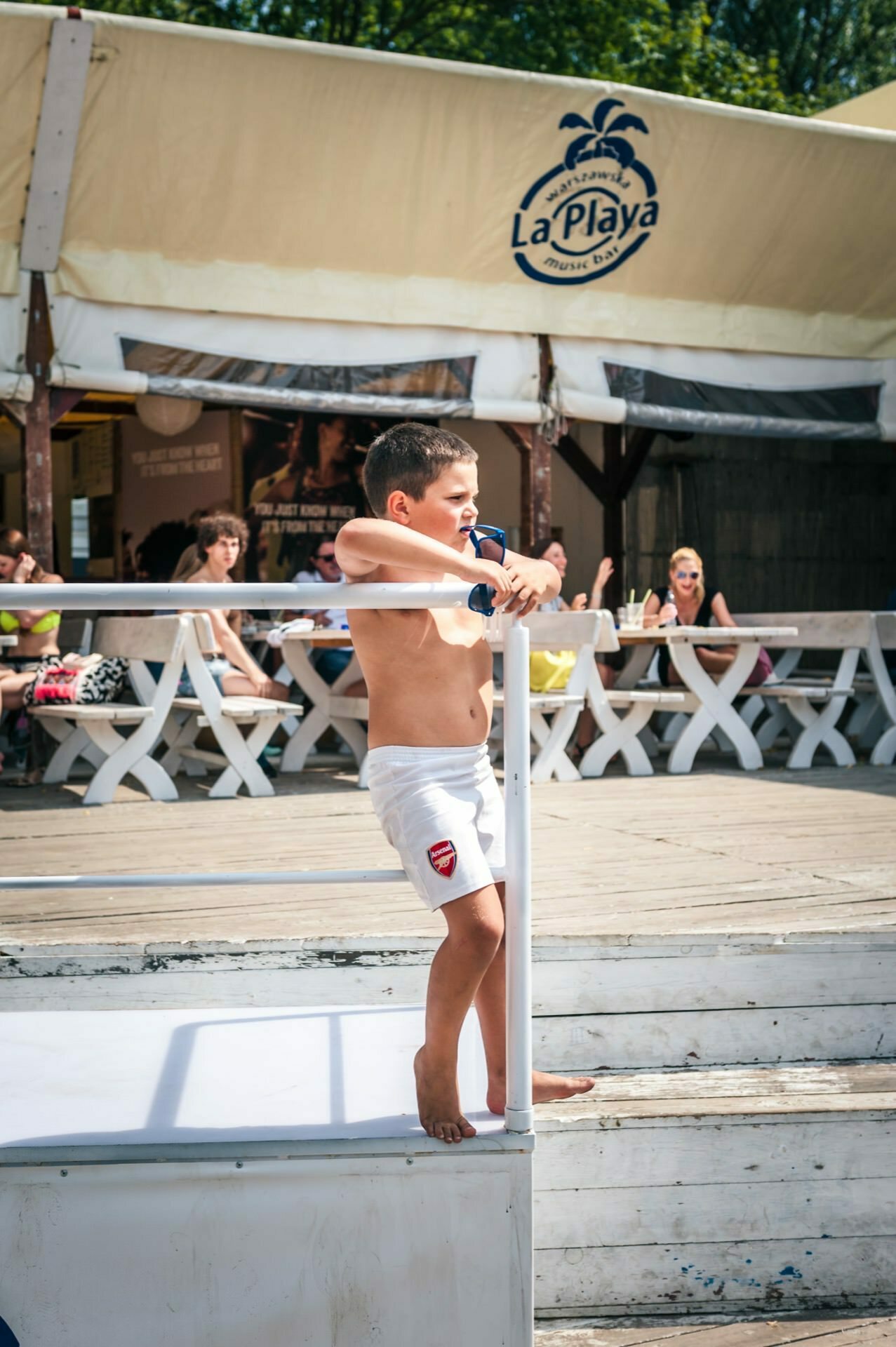 A young boy in white swim trunks stands barefoot on a wooden deck, leaning against a metal railing. Behind him, people sit at white picnic tables under a canopy that reads "La Playa." This tranquil scene could easily be part of a photo essay from the fair showcasing lazy summer moments.  