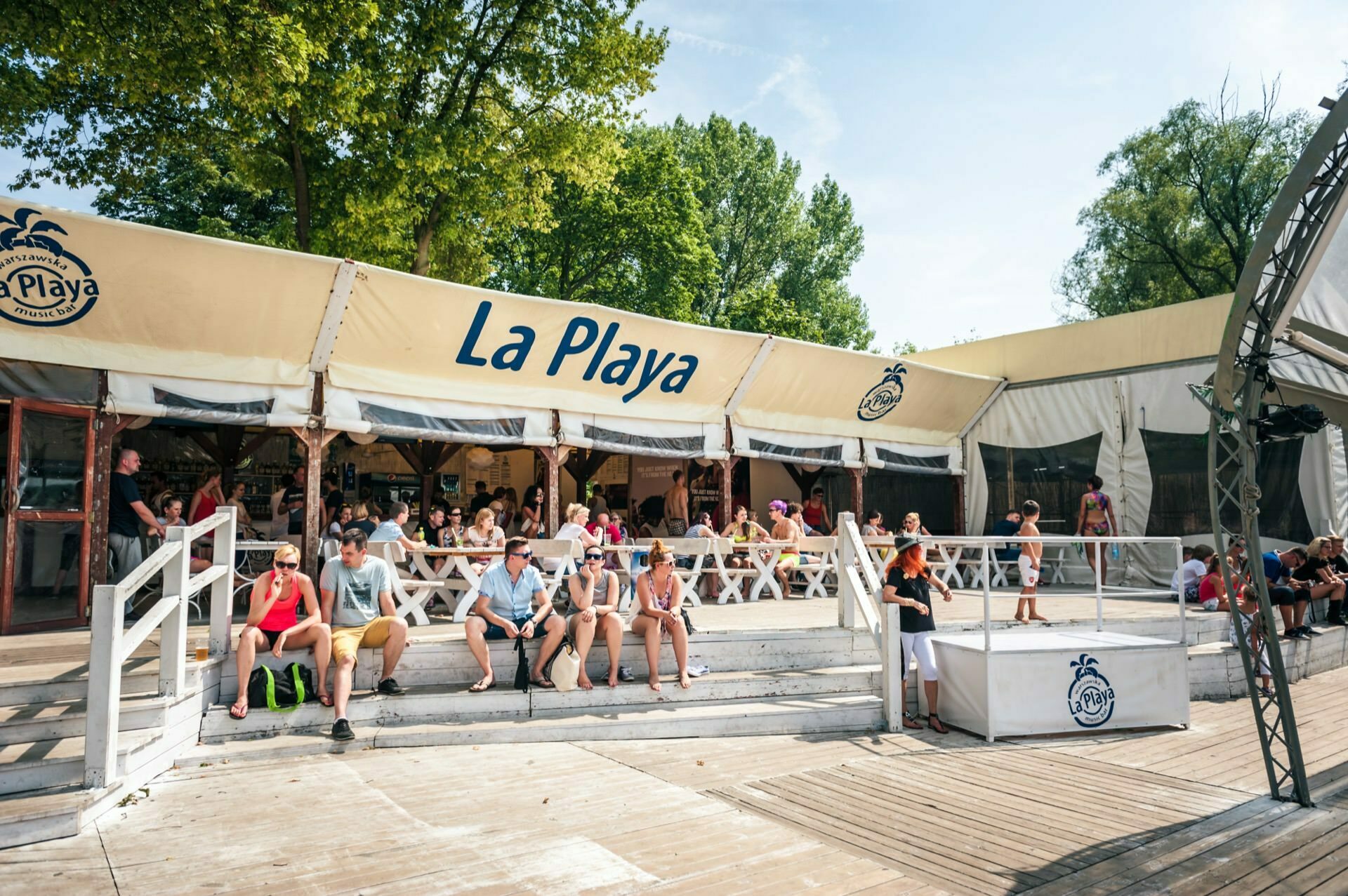 A group of people relax on steps and benches in front of a large, tented establishment called "La Playa." The cream-colored tent, decorated with a prominent logo and name, sets the scene for a sunny day at the event. Trees and blue skies in the background add to the inviting atmosphere captured perfectly in this photo essay of the fair.  