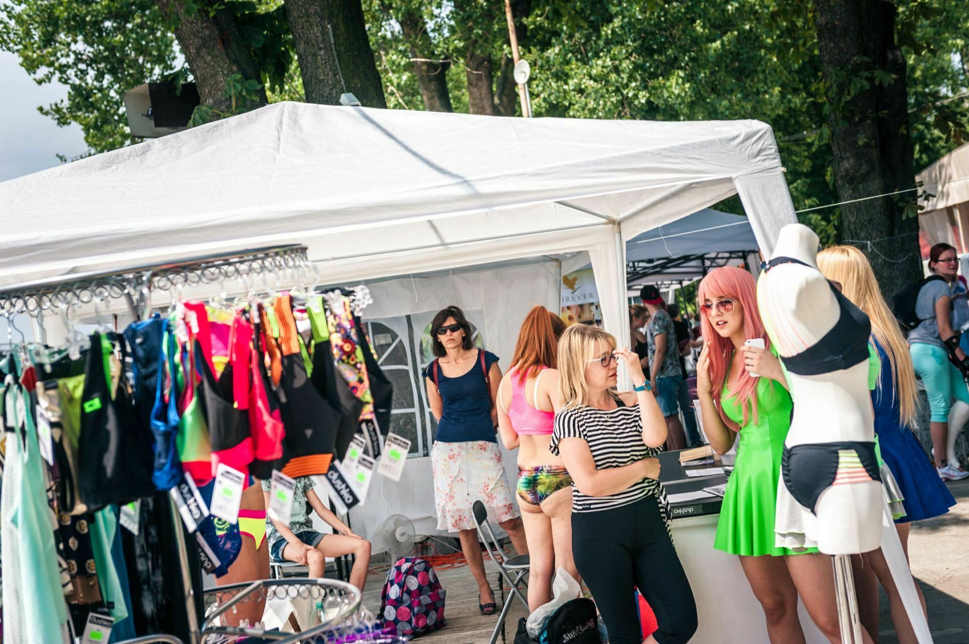 A lively scene at an outdoor market where people browse and chat near a white canopy tent. Colorful swimsuits are displayed on a revolving rack on the left, and a mannequin in a green and black bikini stands on the right. This photo report from the fair shows people enjoying a sunny day.  