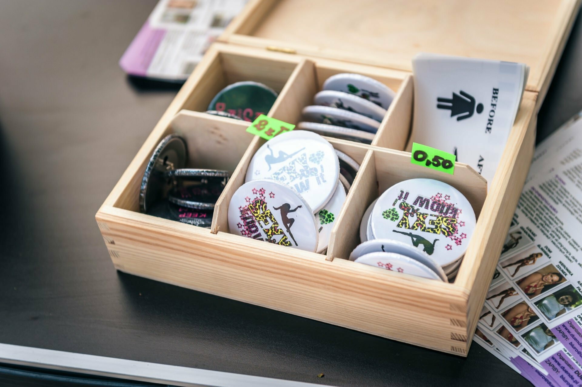 A wooden box containing various round pins with colorful patterns and green prices placed in compartments stands on a dark surface. Some of the buttons are stacked on top of each other, and there are paper replacement symbols and other objects nearby, resembling a photo fair display. 