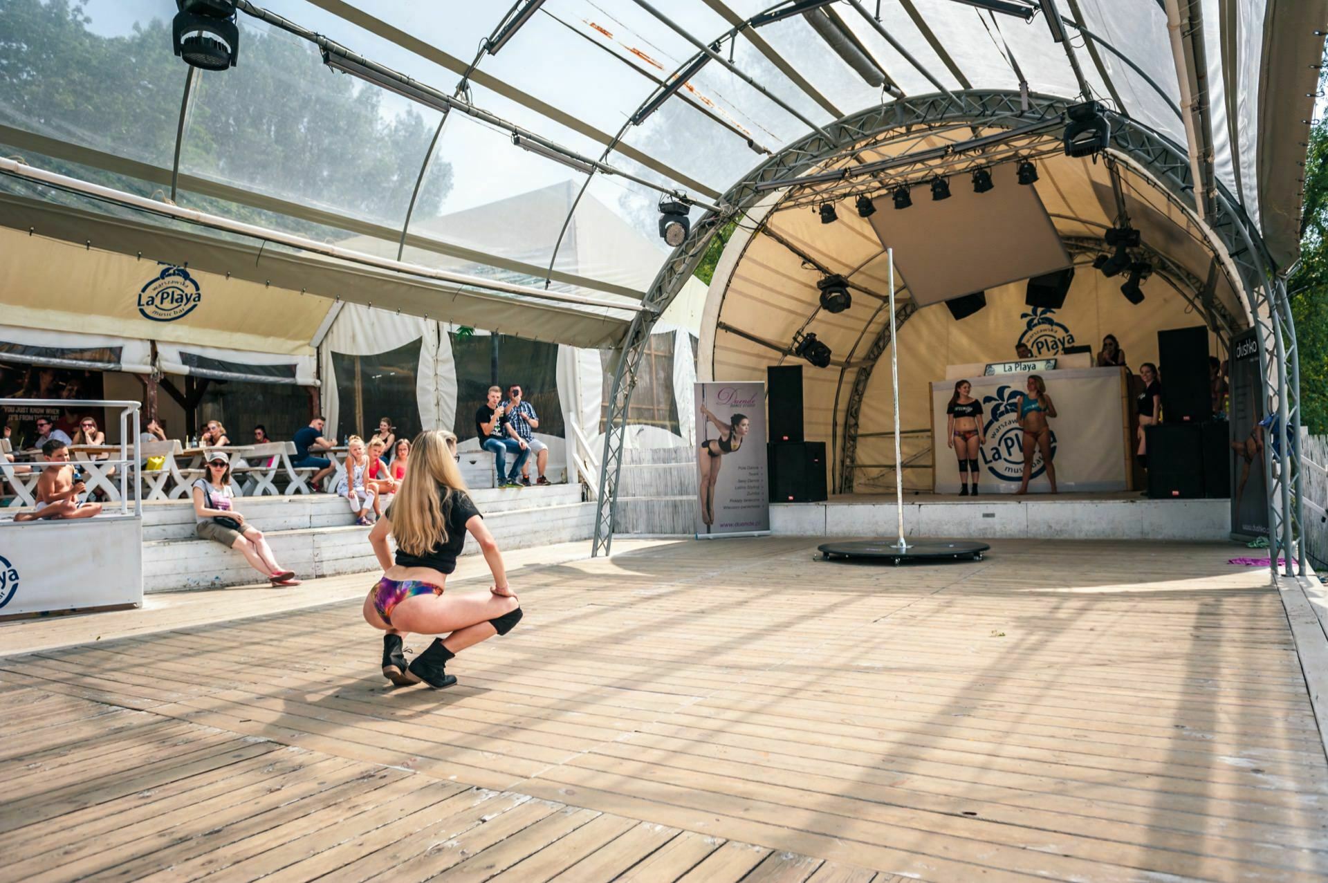 A woman performs a dance move on a large wooden stage under a transparent dome-shaped canopy. Several spectators sit on benches in a shaded area around the stage and watch the performance. A photo essay captured the speakers and a banner with the logo in the background.  