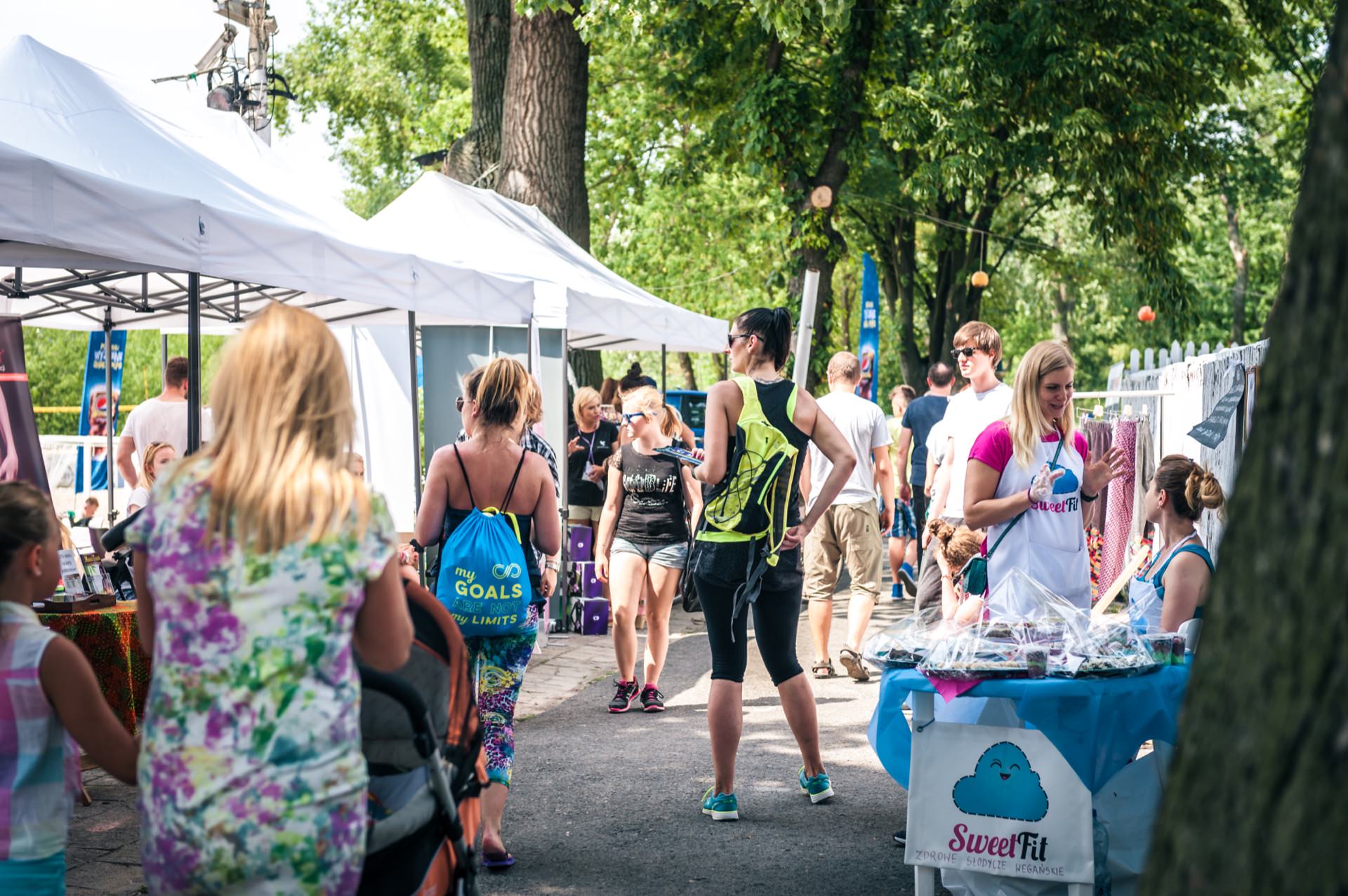 People spend the day outdoors at a bustling market or fair with white tents set up along a tree-lined path. Stalls offer a variety of goods, and a vendor at the "SweetFit" booth interacts with visitors. The atmosphere, perfectly captured in this photo essay of the fair, is casual and filled with bright, sunny weather.  