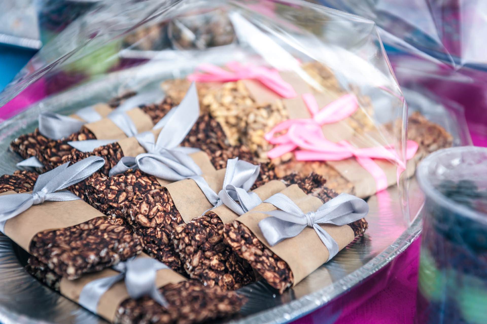 Various homemade granola bars wrapped in brown paper and tied with pink and gray ribbons are displayed on a round tray covered with a transparent lid. In the background are various fuzzy elements that create a colorful setting reminiscent of a vivid photo fair. 