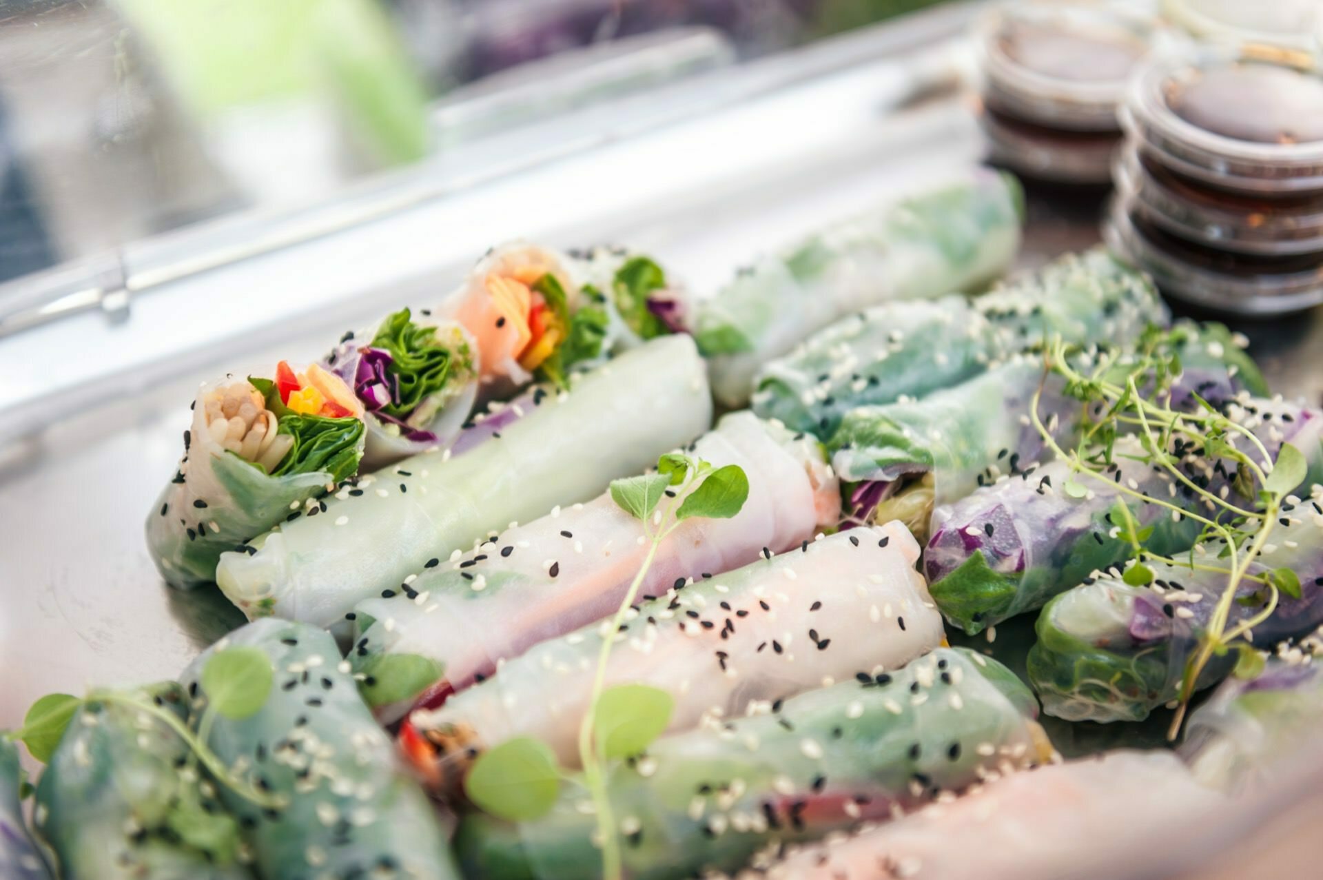 A plate of fresh Vietnamese spring rolls filled with various vegetables, visible through a transparent rice paper wrapper. The rolls, garnished with black sesame and sprouts, are accompanied by small containers of sauce in the background, reminiscent of a delightful exhibition of photo fair food. 