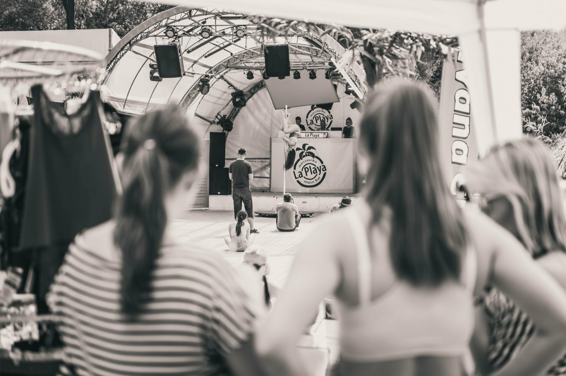 A black-and-white photo showing a photo-op at an outdoor music event. People are sitting on the floor facing the stage with a DJ stand labeled "LaPlaya" under a tent. The foreground is blurred, showing only the backs of three people standing and watching the show.  