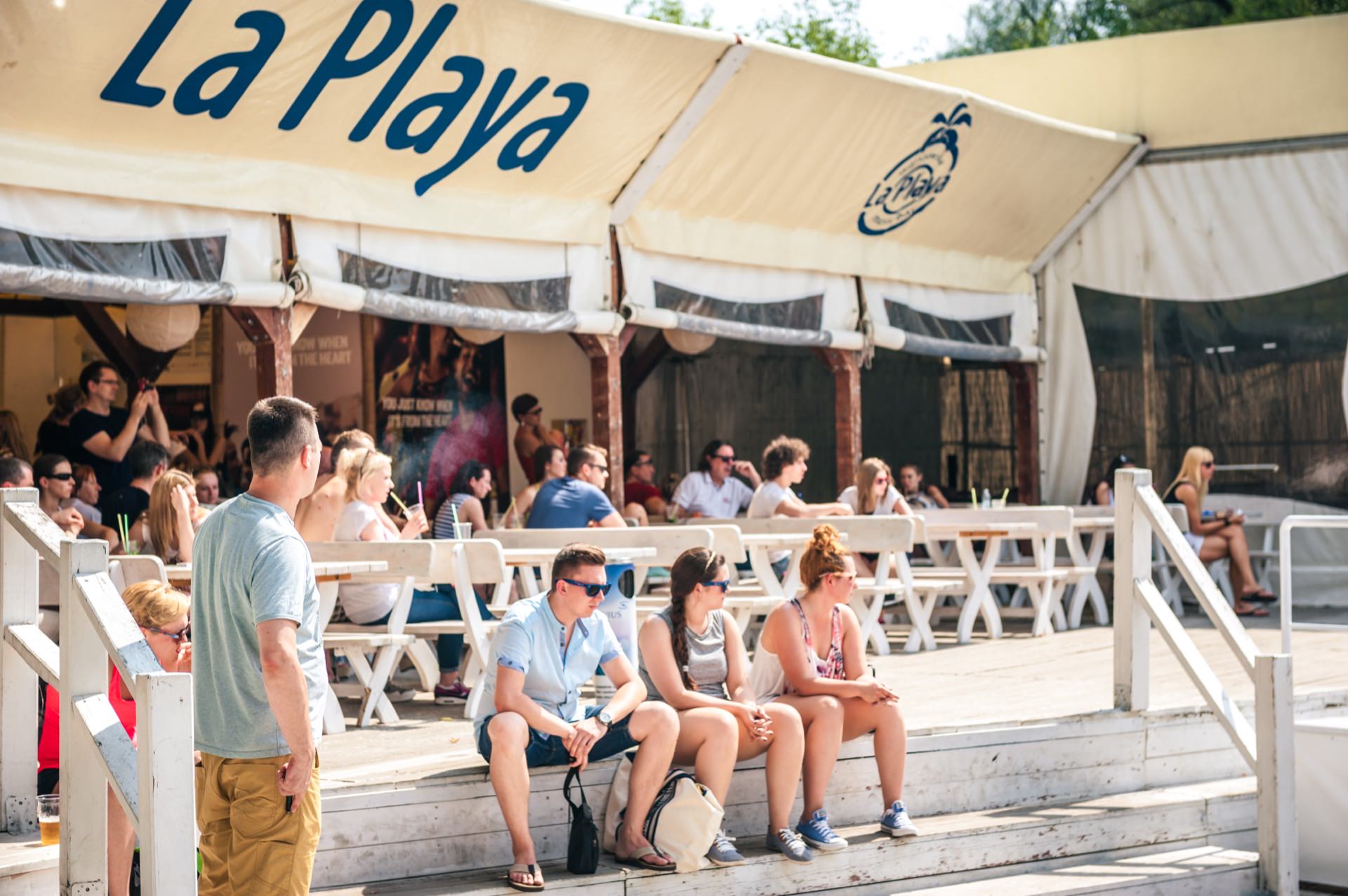 People sit and stand in front of a cafe or restaurant called "La Playa." The area is shaded by a large canopy. Some people sit on white benches, while others stand or walk around in casual summer attire, creating a relaxed and sunny atmosphere. It is reminiscent of a photo fair.   