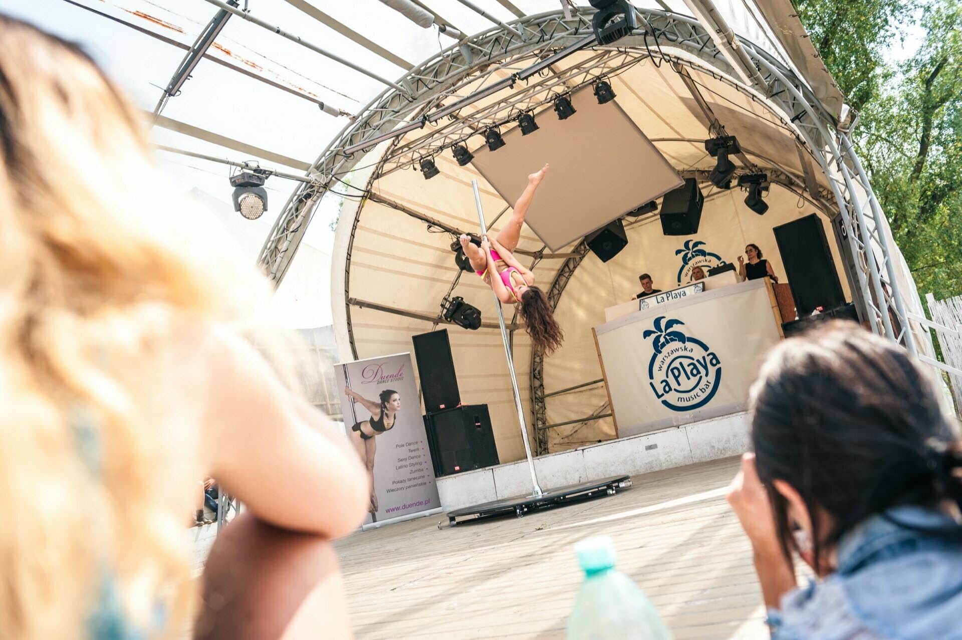 A performer dances on a pole on a stage under a curved canopy. In the background is a DJ booth with the "LaPlaya" logo. In the foreground, the audience can be seen sitting and watching. This bright and sunny scene shows a vibrant photo-op of the fair.   