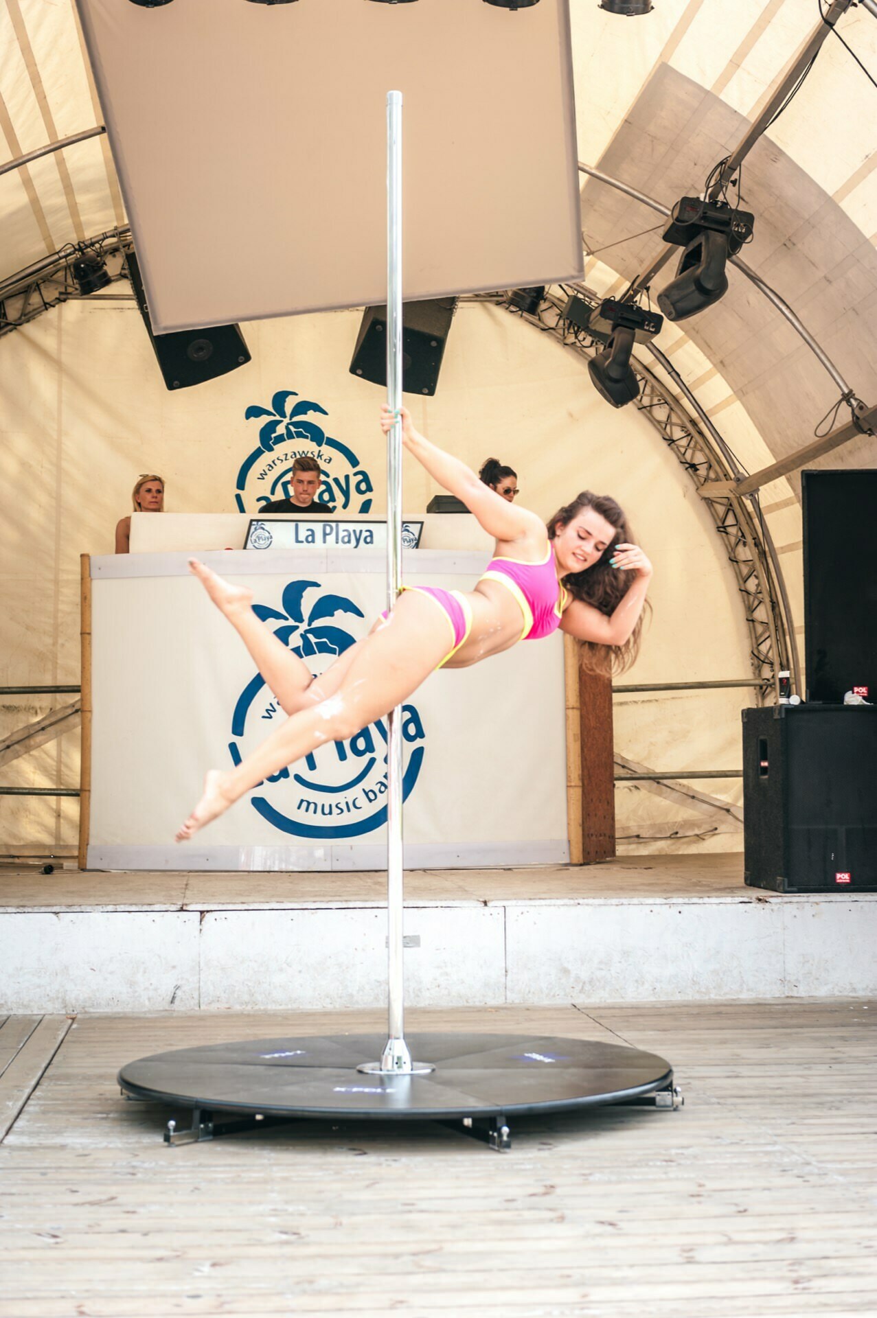 A photo report from the fair captured a woman in a pink top and shorts performing a pole dance on an outdoor stage. Behind her, a DJ is playing music from a booth adorned with the "La Playa" logo. The stage is sheltered by a large tent, while several spectators enjoy the event in the background.  