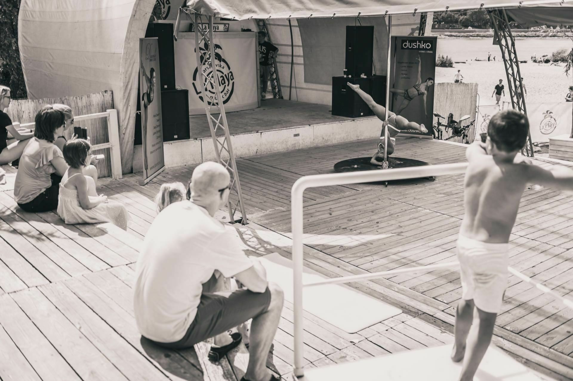 The black-and-white photo serves as a photo essay of the fair, showing an outdoor scene with several people sitting on a wooden terrace. A man and a child are seated on the left and watching intently. Another child is standing by the railing in the foreground, facing an empty stage decorated with props.  