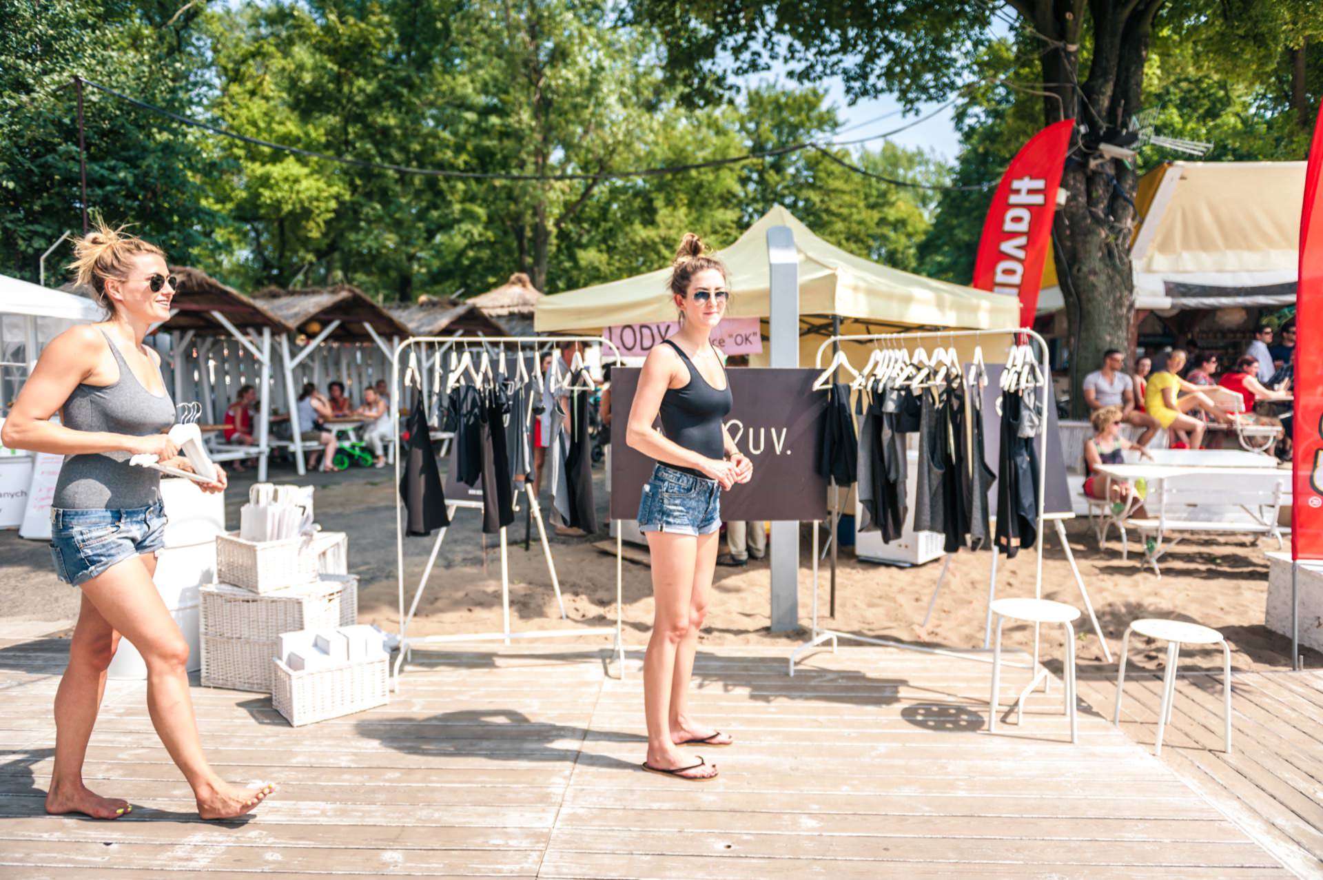 Two women in casual attire, wearing sleeveless shirts and denim shorts stand by a clothes rack at an outdoor market on a sunny day. The market is set up on a wooden terrace with a few stalls and people in the background, all captured perfectly in this photo essay of the market. Green trees provide a lush backdrop.  