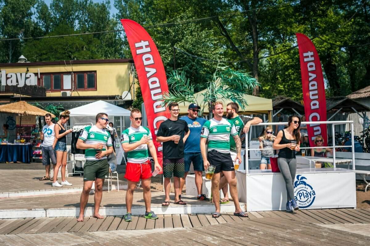 Grupa ludzi stoi na drewnianym pokładzie podczas wydarzenia na świeżym powietrzu. Sześciu mężczyzn w zielono-białych koszulkach sportowych ustawia się w szeregu z przodu, trzech trzyma drinki. Dwa czerwone banery w tle głoszą „HAVANA CLUB”. Fotorelacja z targów pokazuje innych ludzi i widoczne za nimi struktury.