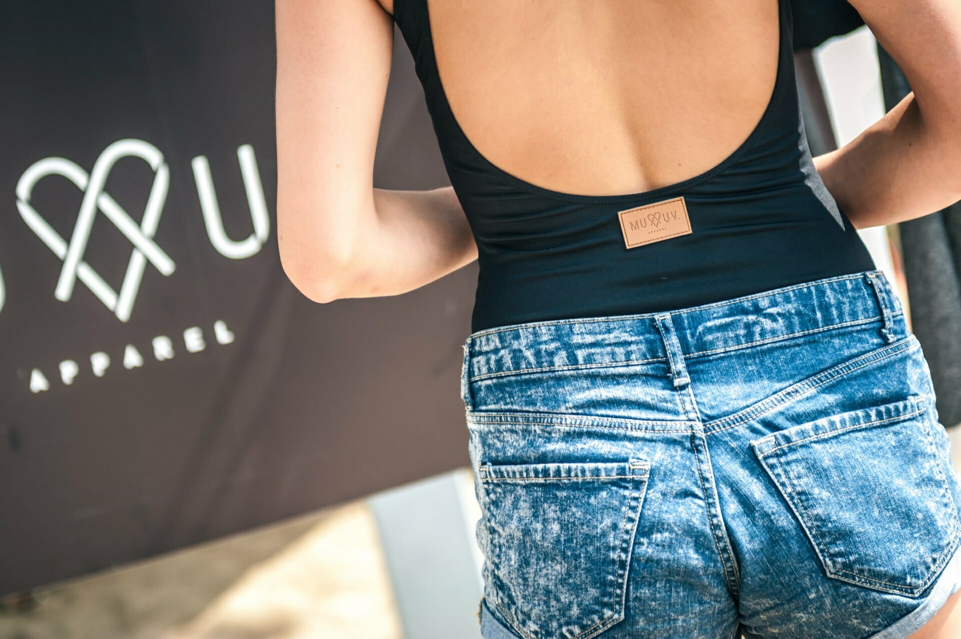 A person wearing a black open-back top and high-waisted denim shorts with rubbed edges can be seen from the back. The top has a small brown tag with the brand name. Part of a sign with a heart and the word "APPAREL" can be seen in the background, adding to the photo fair's atmosphere.  