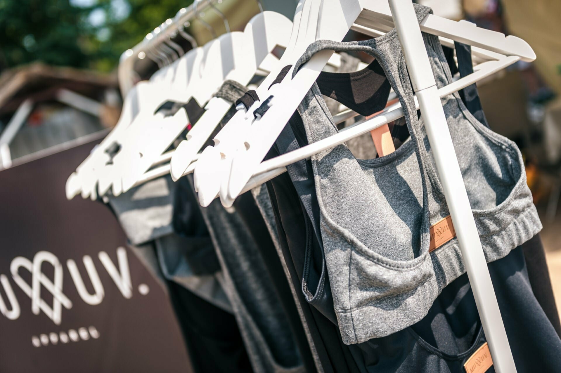 A close-up of several gray sports tops and sports bras hanging on white hangers during a photo tour of the fair. They are displayed on a metal rack outdoors in bright sunlight, with the green visible in the blurry background. 