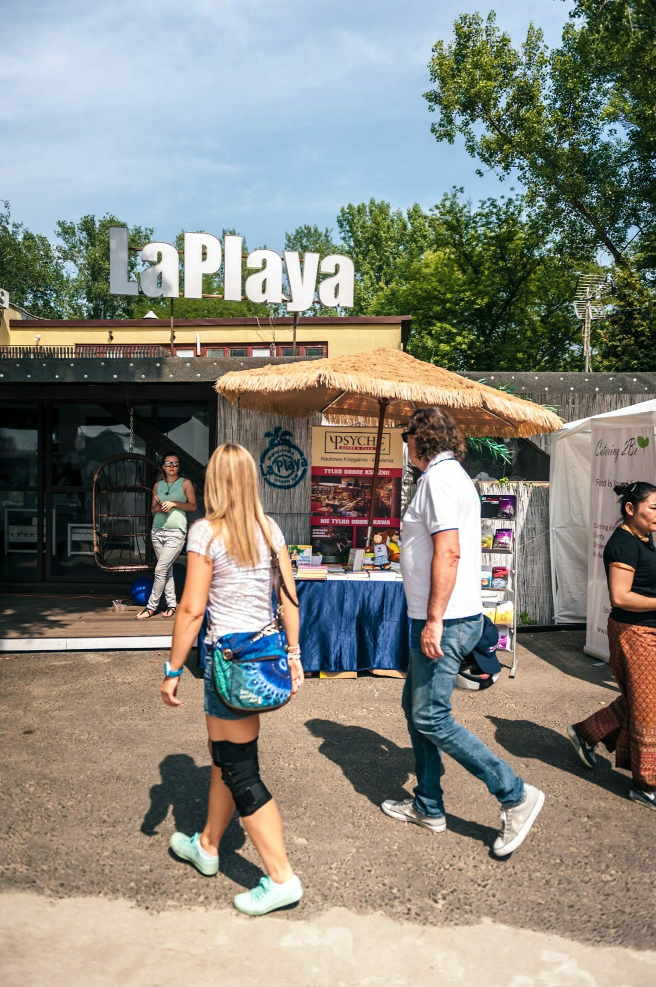 People walk past a vendor's booth at an outdoor market with a "La Playa" sign in the background. The stand, shaded by a thatched roof, displays a variety of products. Trees and other market stalls are visible in the background under a sunny sky, creating an idyllic scene perfect for a photo-op of the fair.  