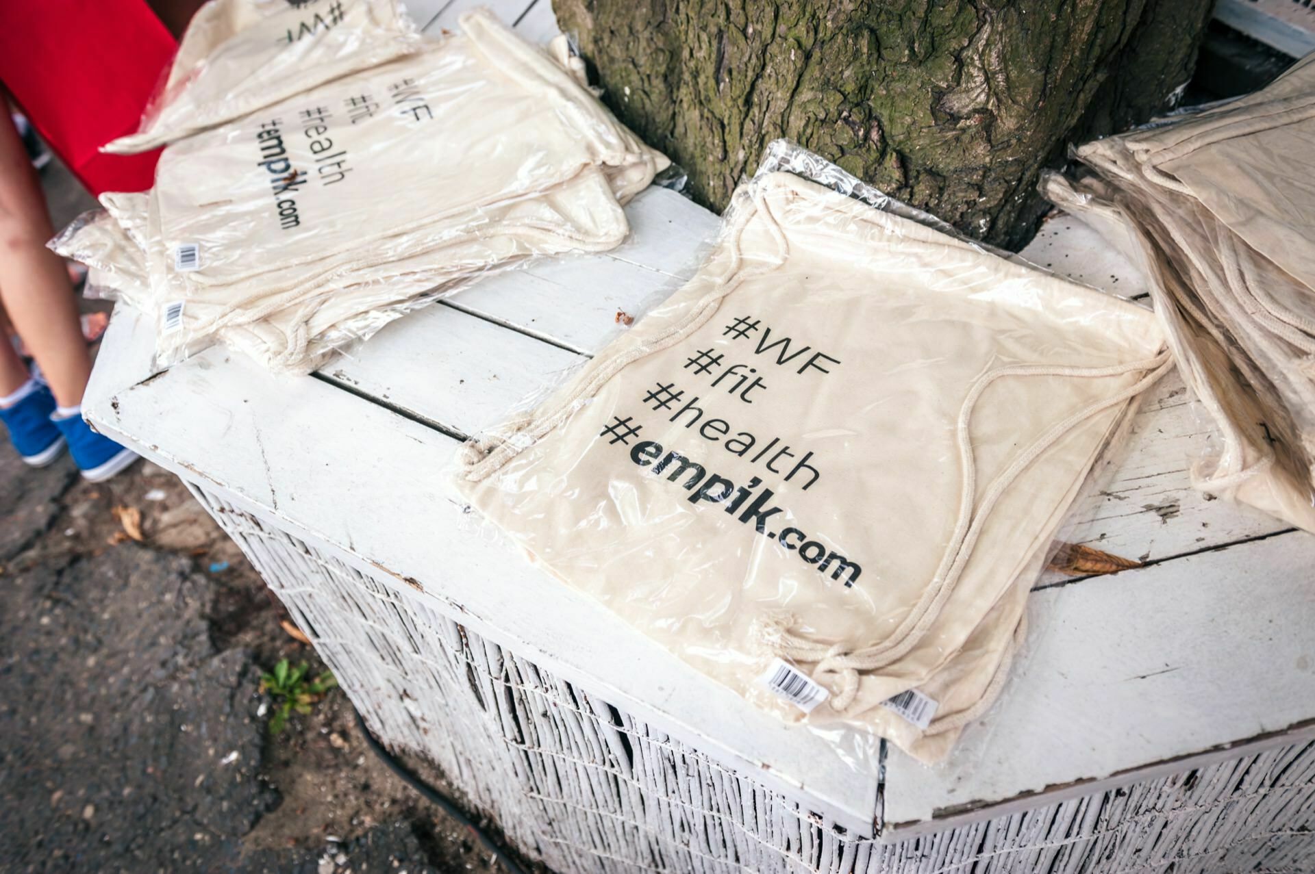 Wrapped in plastic, the beige T-shirts printed with the hashtags #WF, #fit, #health and empik.com are displayed on a white wooden table under a tree. The photo report from the fair captures the essence of these trendy items in their natural setting. 