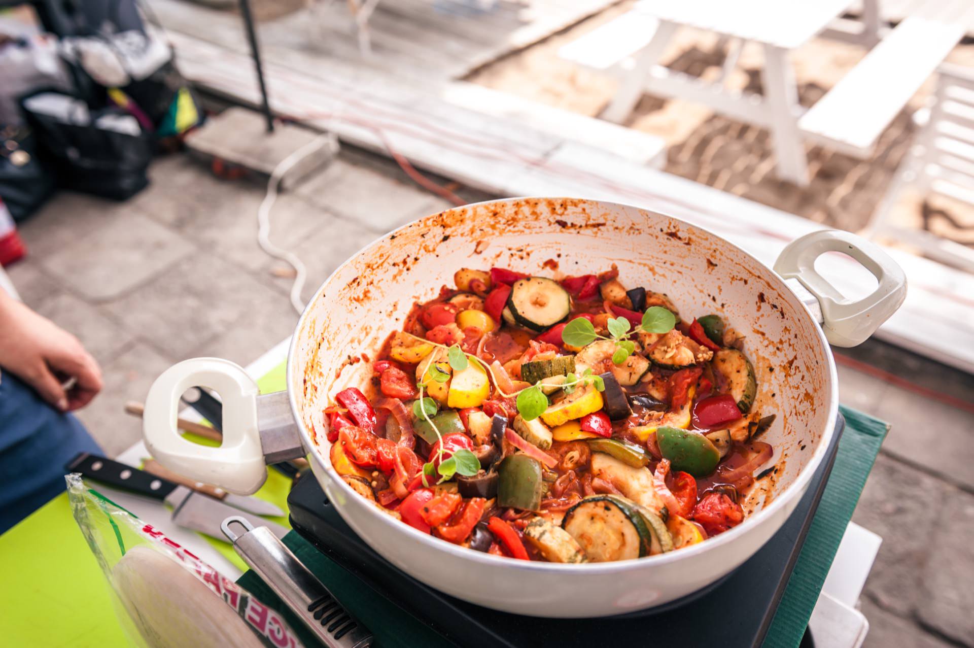 A white ceramic pot filled with colorful vegetable stew, including peppers, zucchini and herbs, stands on a green surface. In the background are a blur of picnic tables and various items from our photo essay of the fair. 