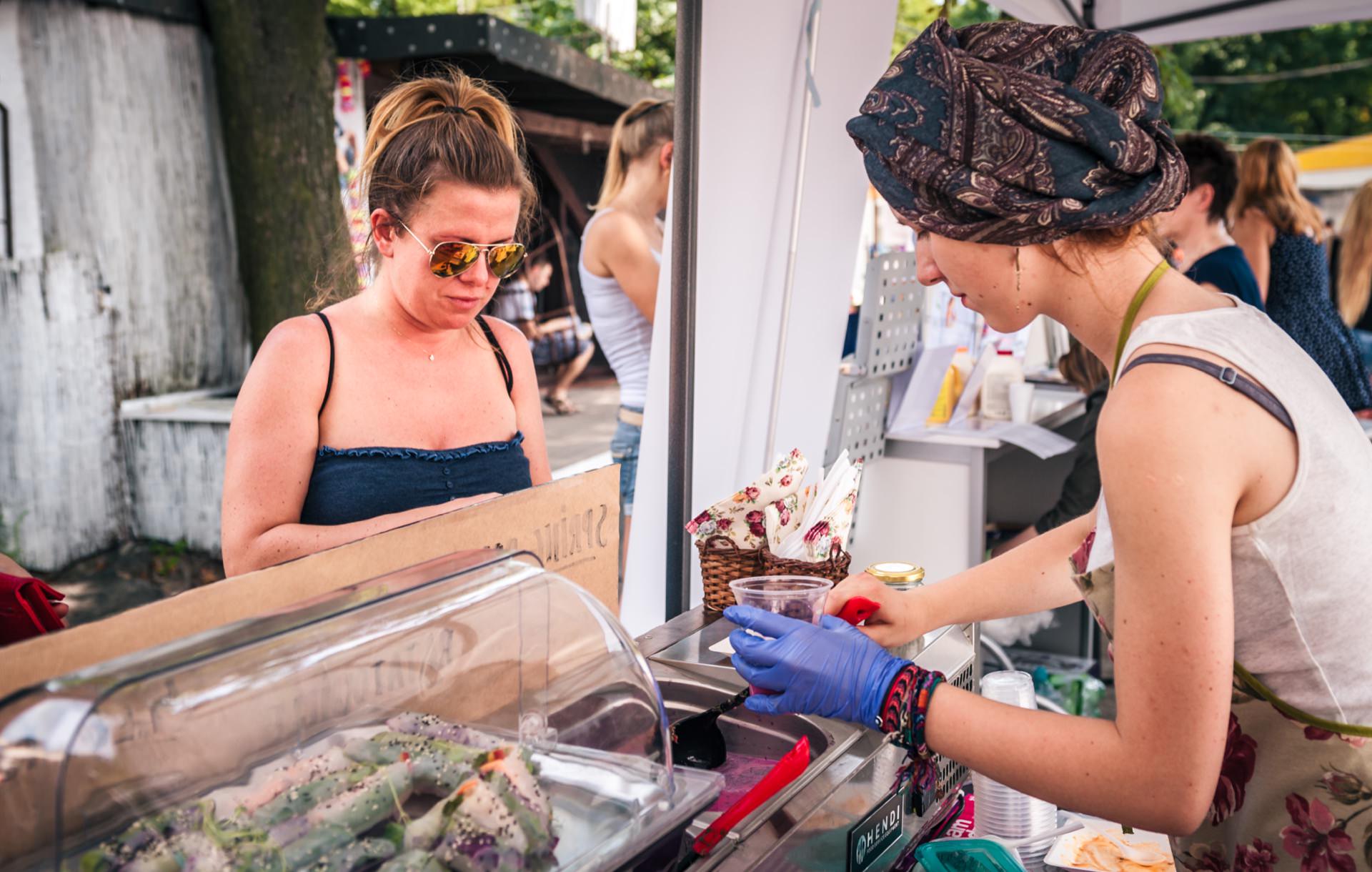 A woman wearing sunglasses is browsing the food offerings at a stand at an outdoor market with a vendor wearing a patterned head scarf and gloves. The vendor is preparing a dish, and various food products are displayed under a transparent cover on the counter, creating a lively scene perfect for a photo-op of the market. 