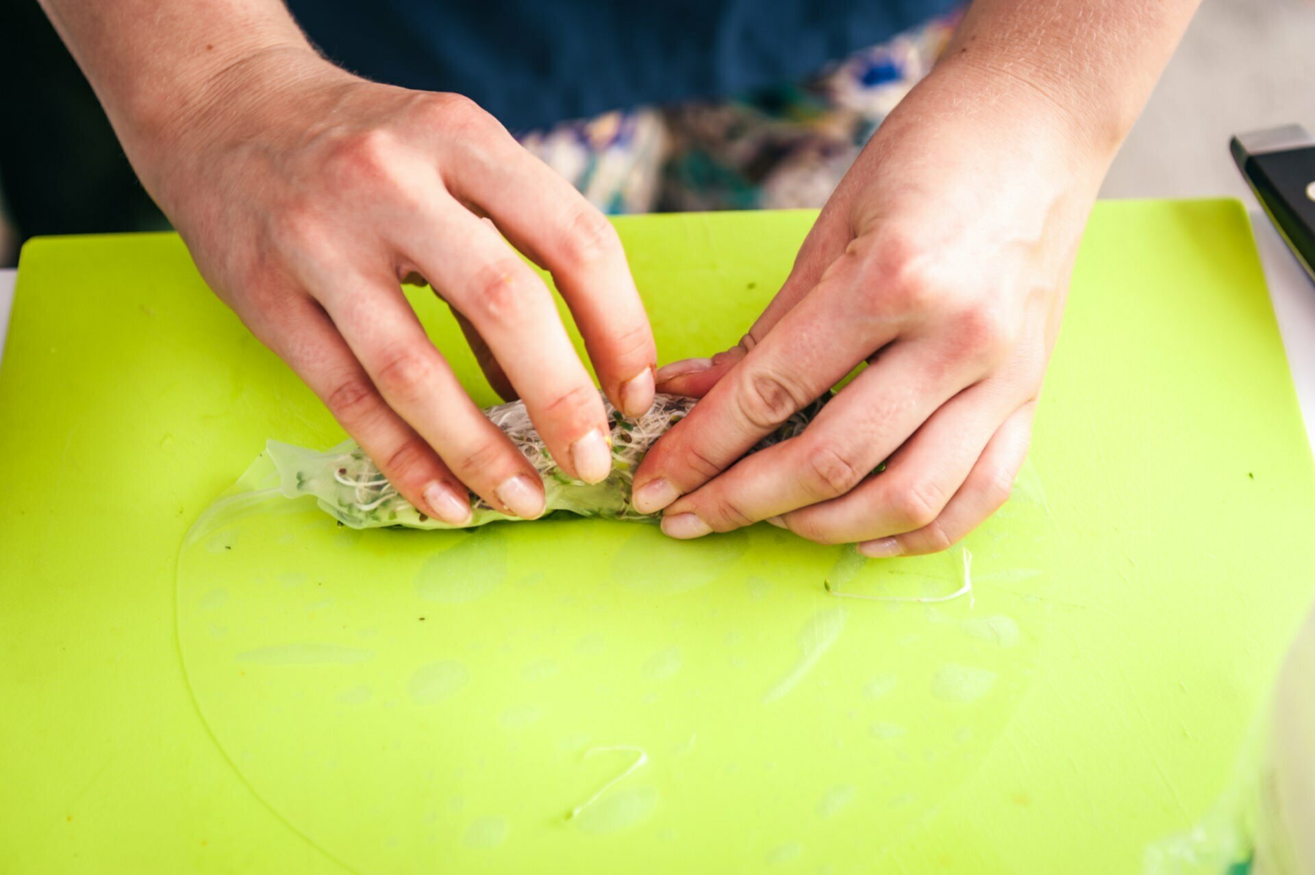 Close-up of a pair of hands rolling up a summer roll on a bright green cutting board, reminiscent of a photo-report from a fair. The person appears to be wrapping the shredded ingredients in thin rice paper. The cutting board and ingredients are slightly wet.  