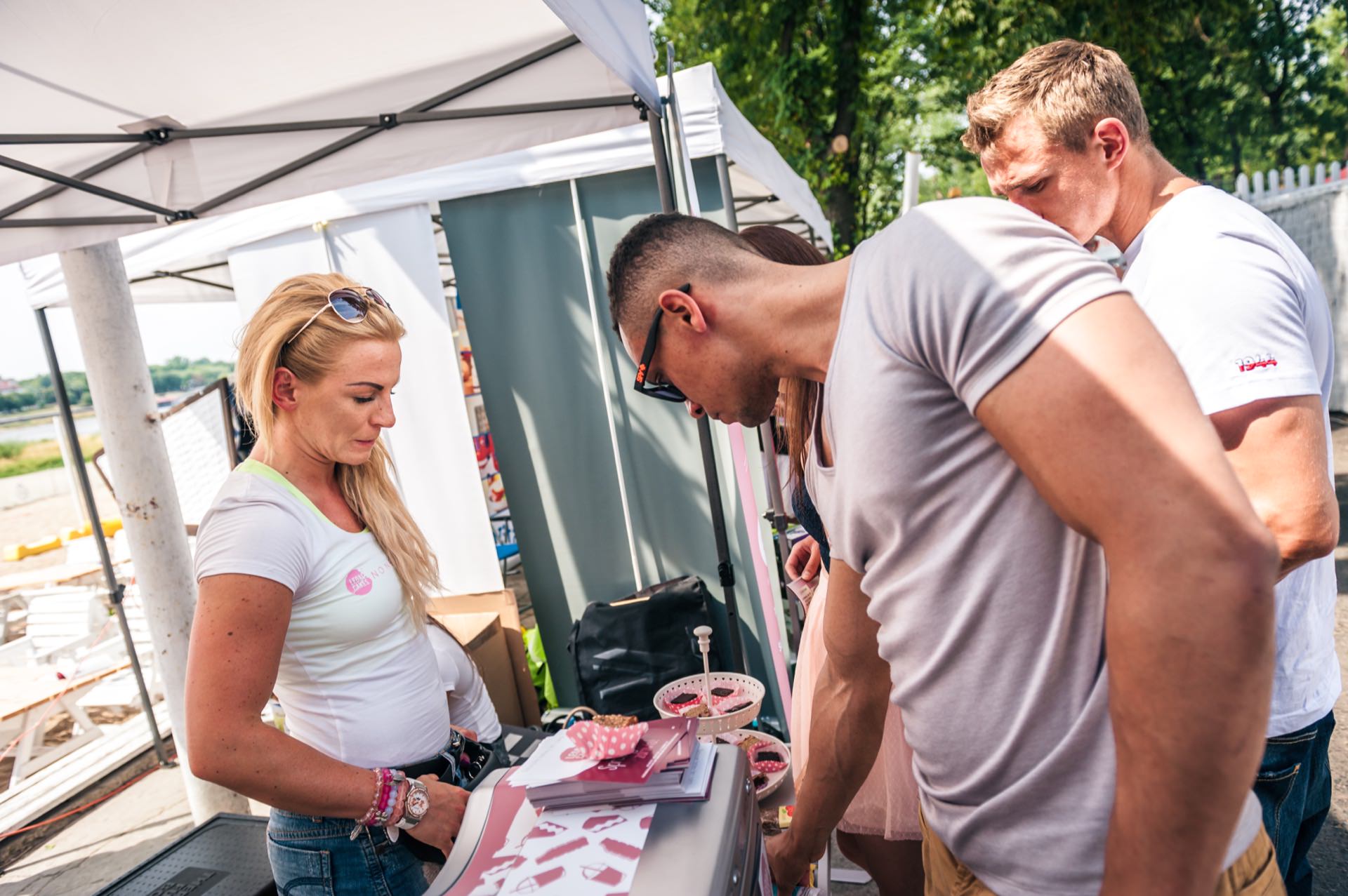 A group of people are interacting at an outdoor booth under a canopy. A woman with blonde hair stands behind the booth while two men look at the items on display. The sunny setting suggests that this is an event or fair. The photo report of the fair perfectly captures the lively atmosphere.   