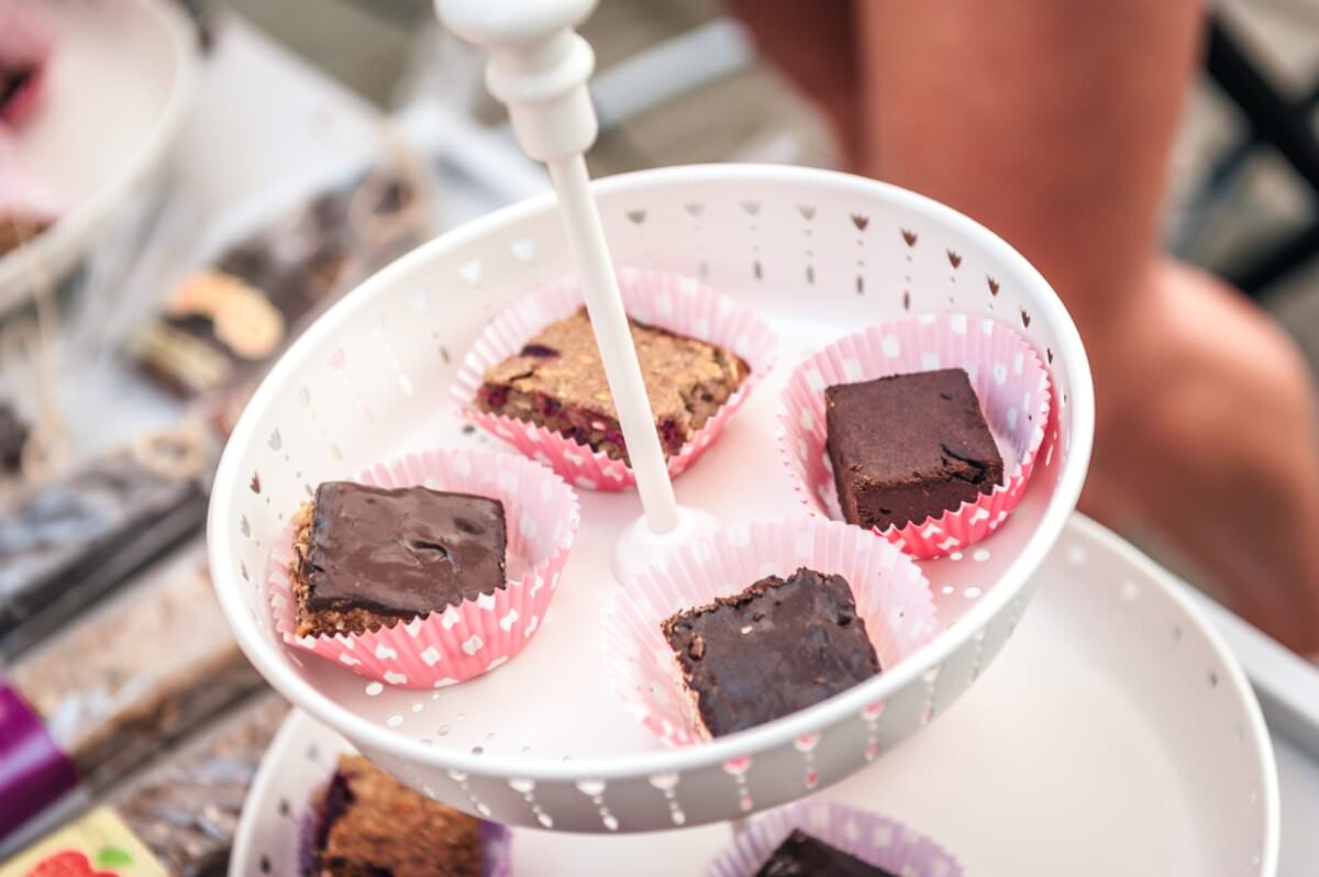 Zbliżenie na dwupoziomową białą metalową tacę do serwowania z różowymi papierowymi wkładkami w kropki, na której znajdują się różne brownie i batony. Taca jest wystawiona na stole na zewnątrz podczas wydarzenia Fotorelacja z targów, a w tle widoczne są inne wypieki.