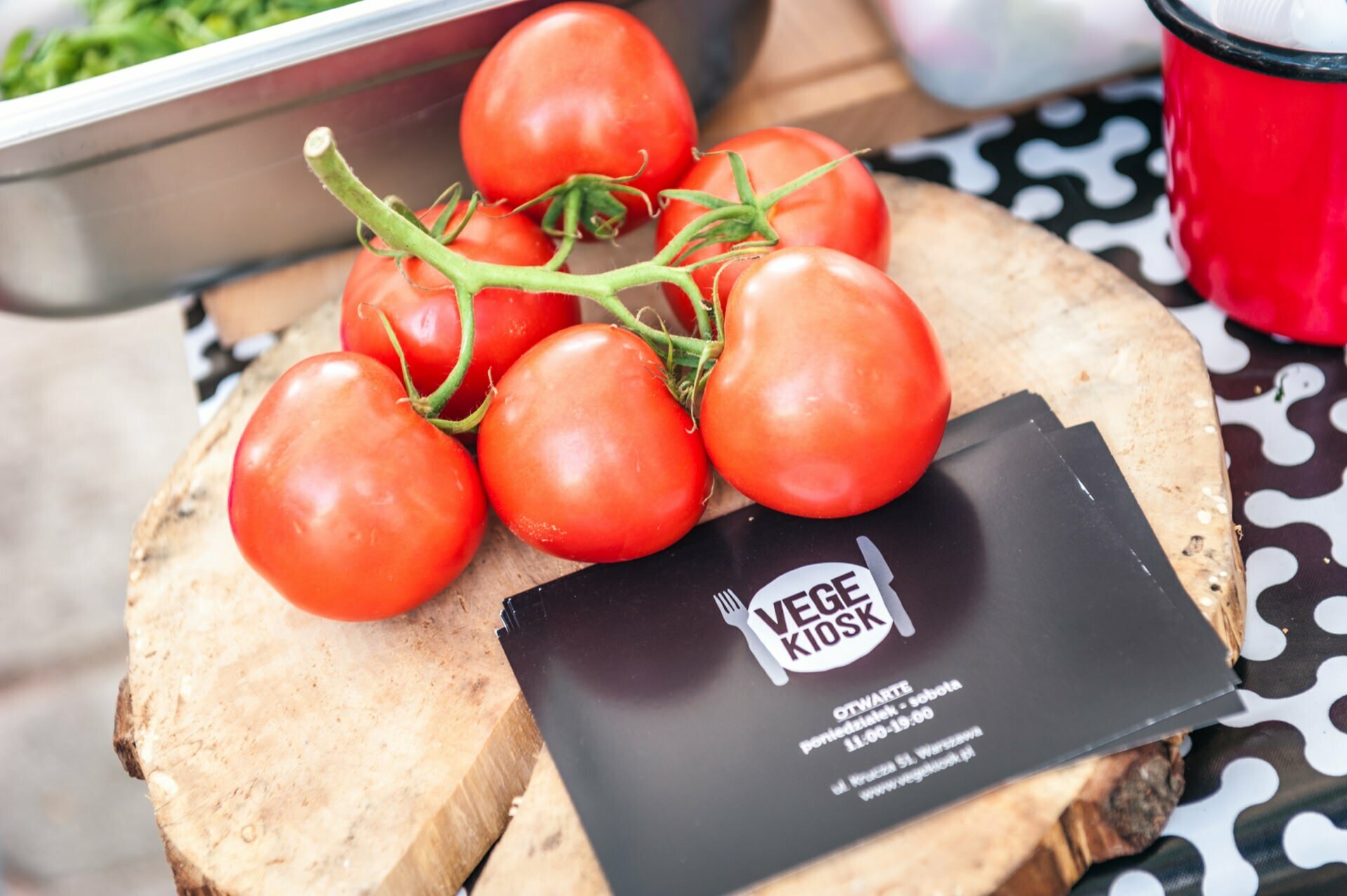 A cluster of ripe tomatoes lies on a wooden cutting board next to a stack of black "Vege Kiosk" business cards. The installation, captured in a photo essay from the fair, is part of an open-air trade show display, with a container of fresh greens visible in the background. 