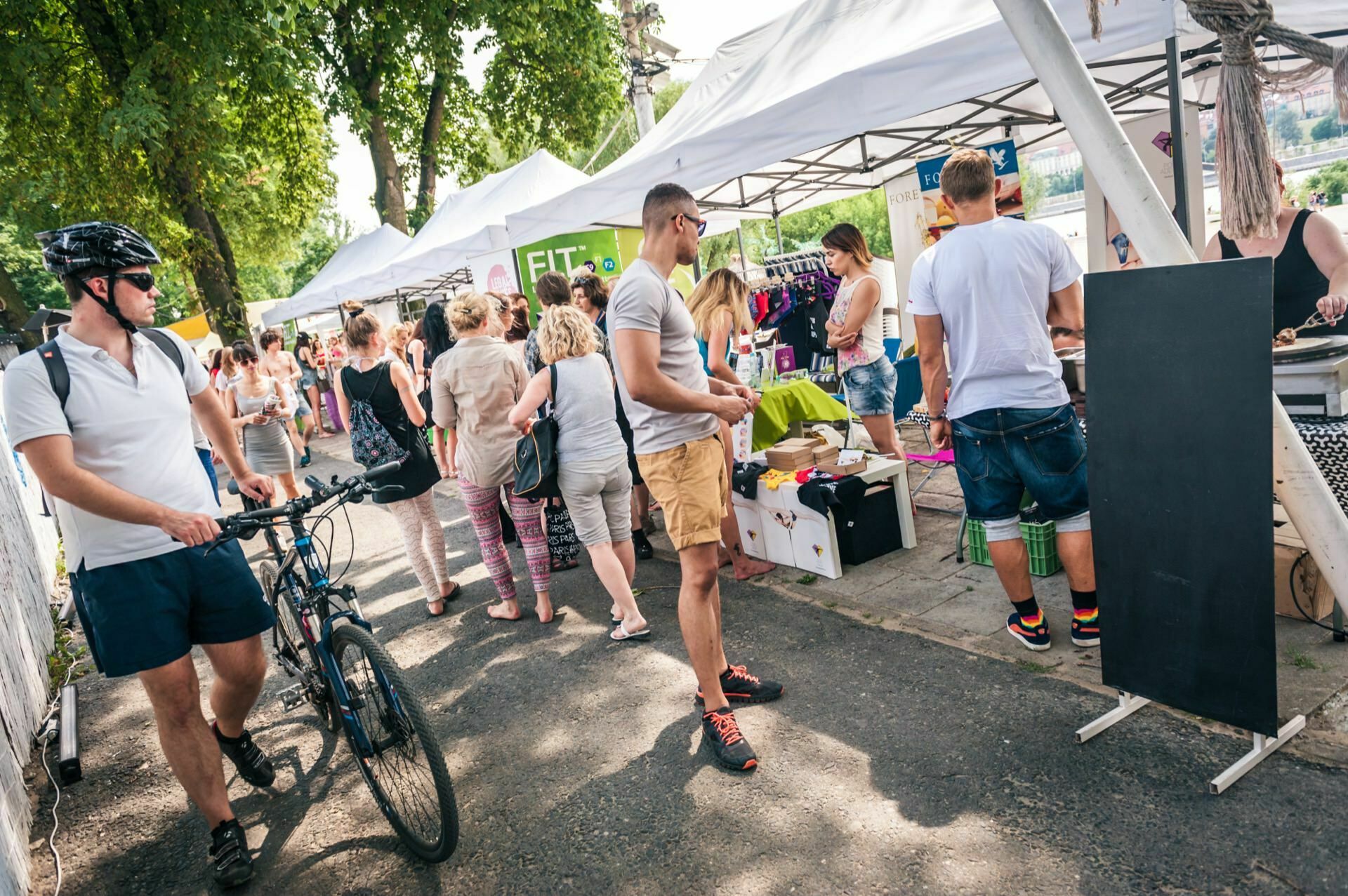 People are enjoying the outdoor market on a sunny day. Many are browsing the various stalls with tents that are set up along the path, captured in a photo essay of the fair. One person is riding a bicycle, while others are looking at items for sale. Trees in the background provide some shade.   