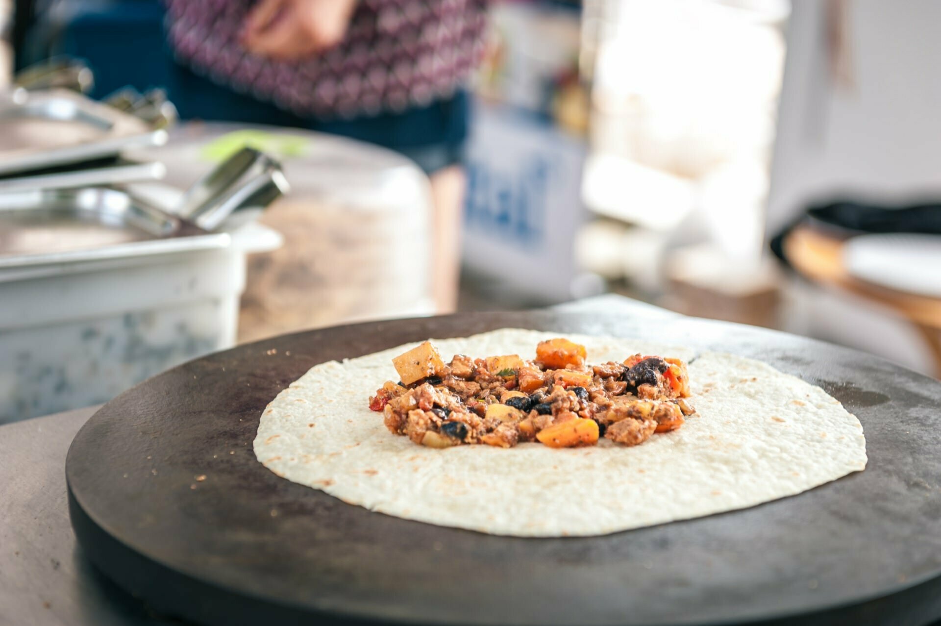 Close-up of a tortilla on a flat cooking surface, with a mixture of diced vegetables and meat. The background is blurred, revealing hints of a photo fair with a person in a patterned shirt and some cooking utensils. 