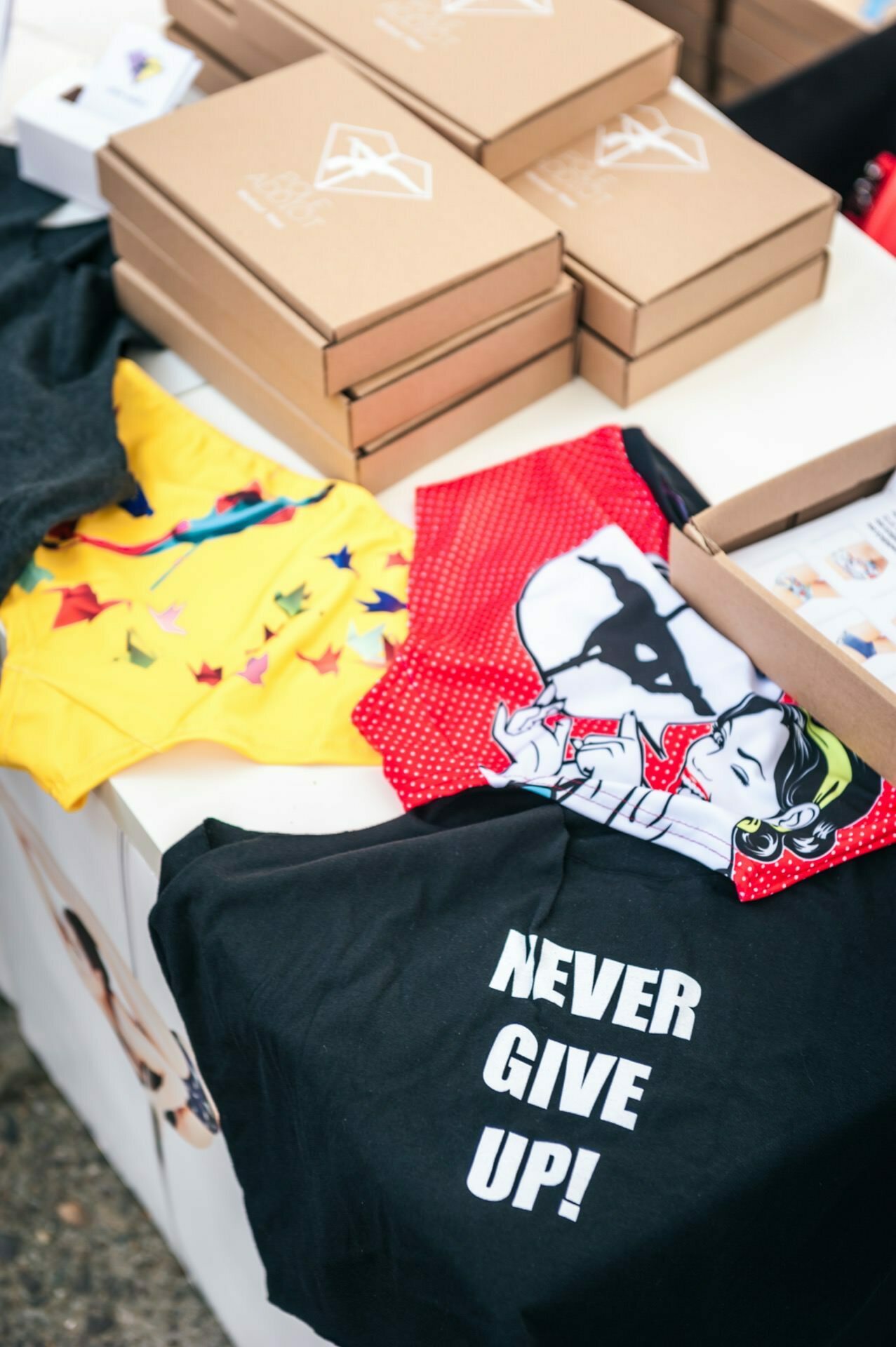 A table of various colored T-shirts on display, captured in our photo report from the fair. One black T-shirt proudly bears the words "NEVER GIVE UP!" and a red polka dot T-shirt has a graphic depicting a man. The brown cardboard boxes are neatly stacked on a table.