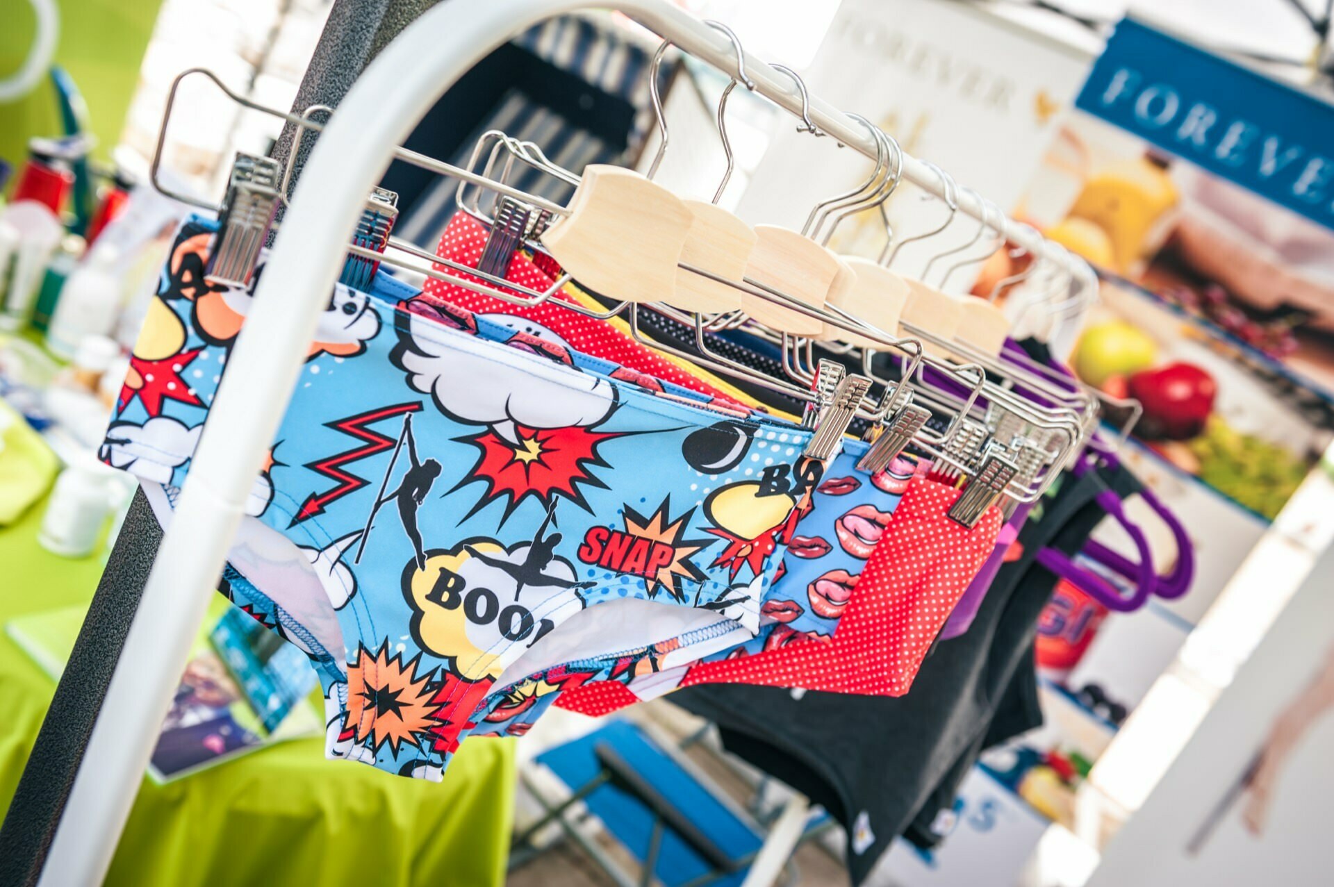 The stand holds several pairs of colorful underwear hung on wooden hangers. The front pair has a cartoonish design with words like "BAM," "SNAP" and "BOOM" next to a cartoon graphic. A background photo reveals the busy, brightly lit interior of the market.  