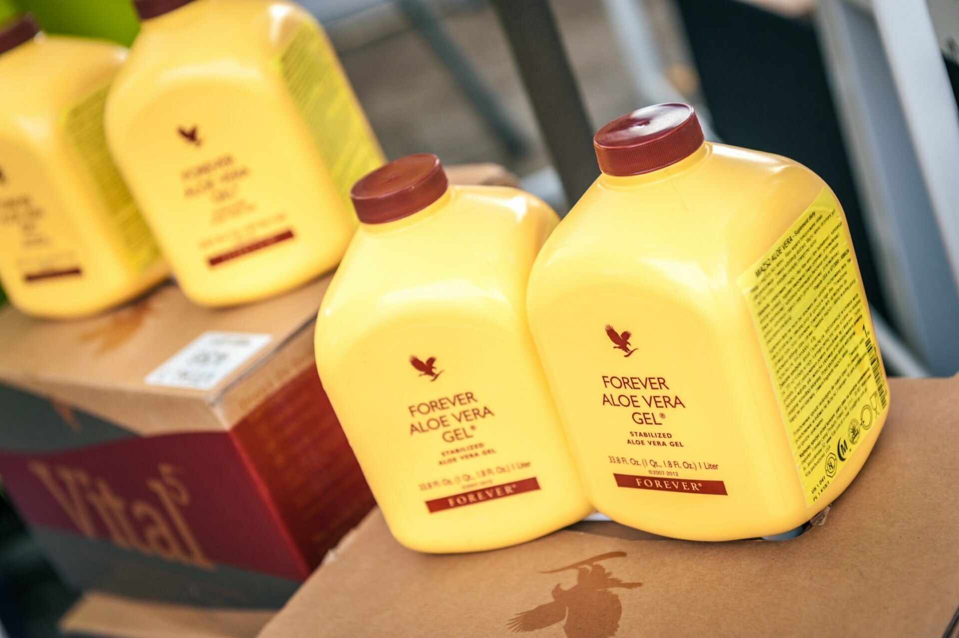 Several yellow bottles of Forever Aloe Vera Gel, each with a maroon lid and maroon logo and text, set on a cardboard box. The background shows more bottles and blurry objects, capturing a snapshot from our photo tour of the fair. 