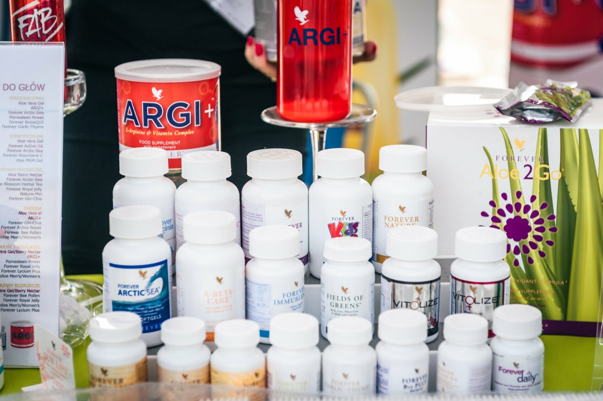 Various bottles of health supplements lined up on the table, including products such as ARGI+, Forever ARCTIC SEA, Forever Calcium and Forever Immublend. In the background is a tall glass with a red drink labeled ARGI+. The scenery appears to be an exhibit or display photographed at the fair.  