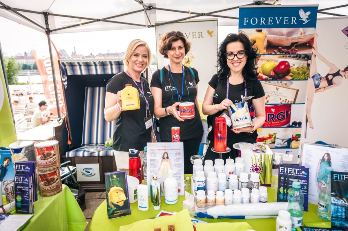 Trzy kobiety uśmiechają się stojąc za stoiskiem wystawowym prezentującym różne produkty zdrowotne i wellness marki „Forever”. Stoisko, zaprezentowane w tej fotorelacji z targów, obejmuje butelki, broszury i plakaty z informacjami o produktach i marką. Znajdują się na zewnątrz, w tle widać budynki miejskie.