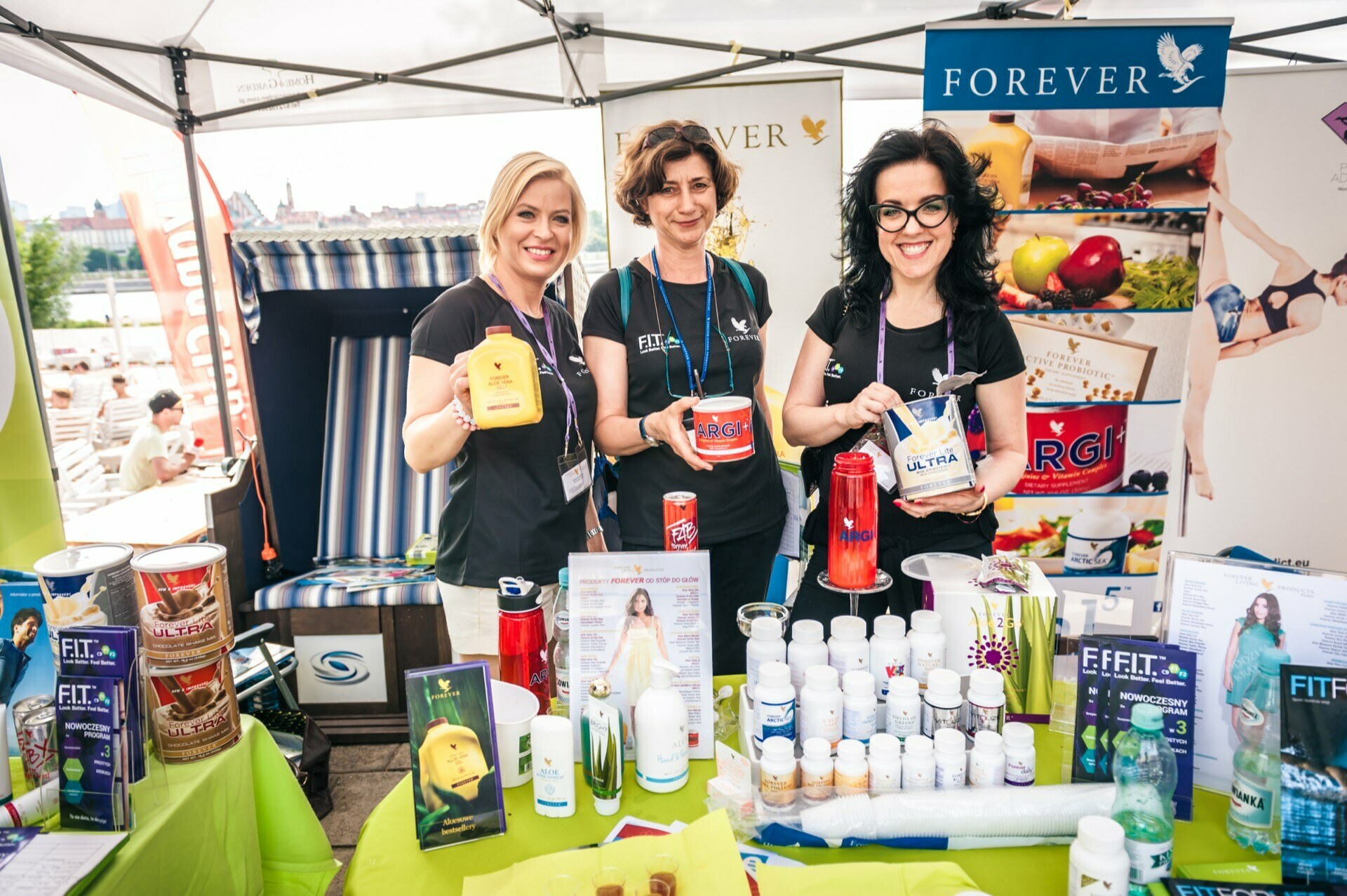 Three women smile as they stand behind an exhibition booth displaying various health and wellness products of the "Forever" brand. The booth, shown in this photo essay from the fair, includes bottles, brochures and posters with information about the products and the brand. They are located outside, with city buildings visible in the background.  