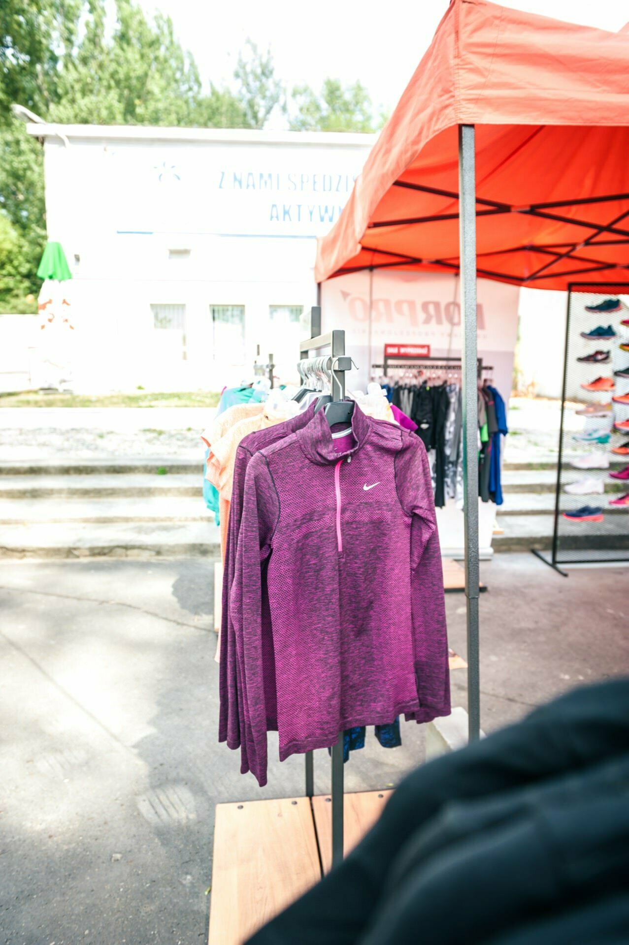 Photo report from the fair: A clothing display under an orange canopy at an outdoor market. The main focus is on a purple zippered jacket hanging on a hanger, while various colorful clothes and shoes are visible in the background. 