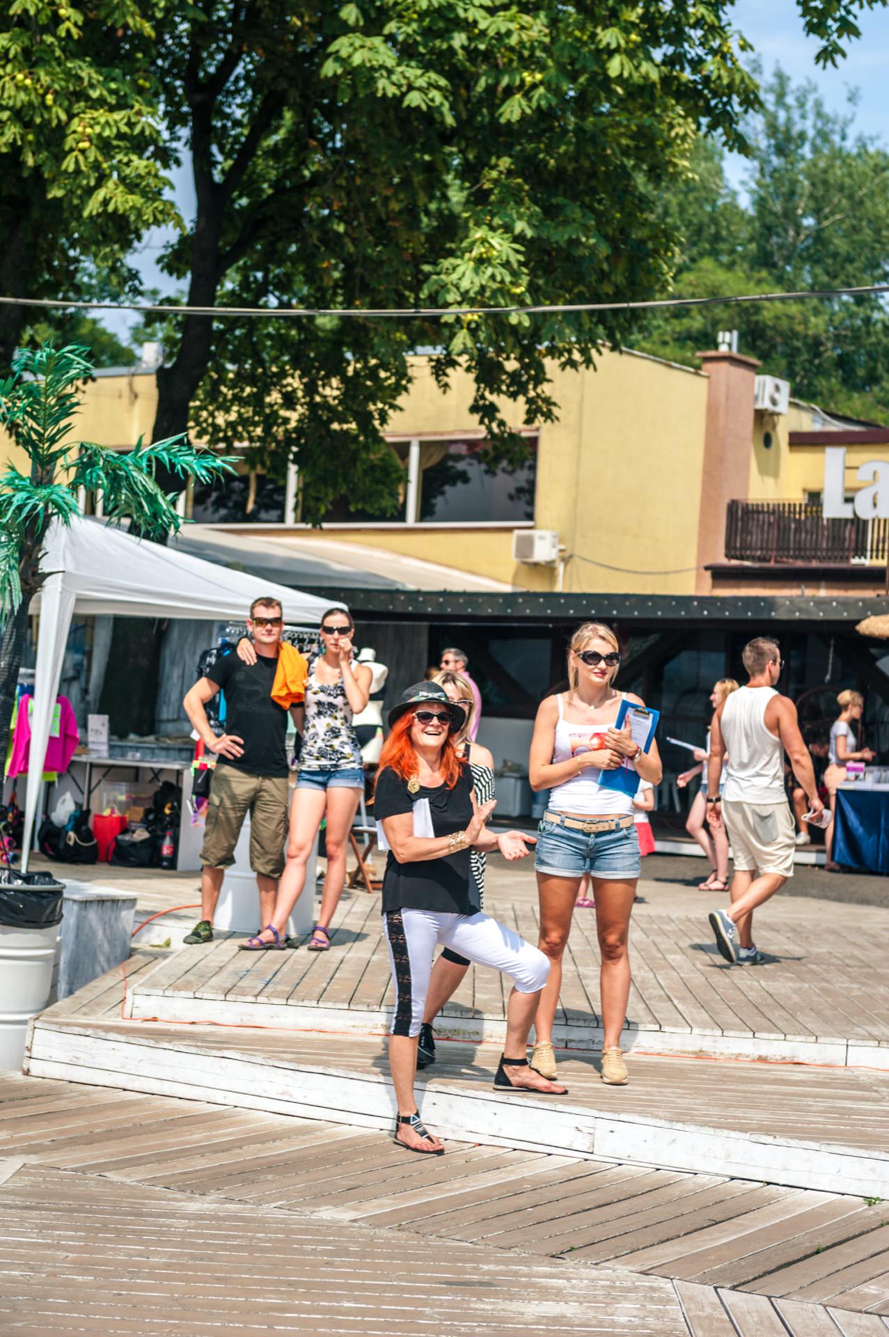 A group of people enjoying a sunny day at an outdoor event. Some people are standing on a wooden terrace, and one woman in front is posing and smiling. The people in the background seem relaxed and engaged. Trees and a building are visible in the background, completing the photo fair.   