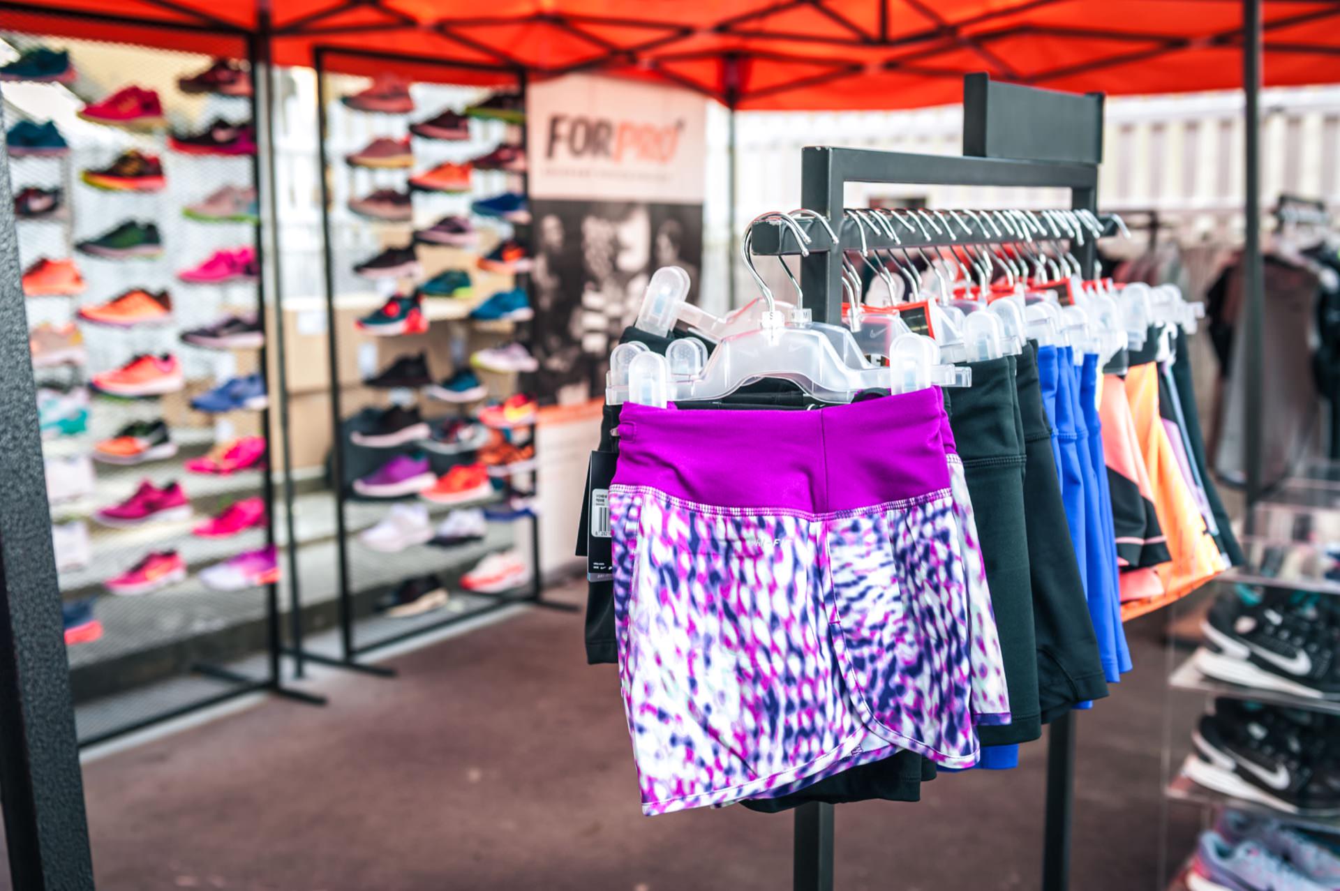 In the foreground of a stall that is part of a vibrant photo fair, colorful sports shorts are hung on hangers. There is a backdrop of various sports shoes on the shelves, while a bright red canopy and sports-related posters create an energetic atmosphere in the background. 