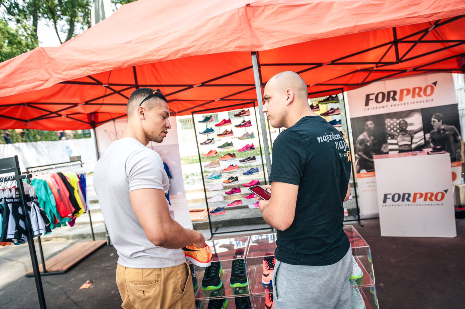 Two men stand under an orange tent, looking at sports shoes spread out on a table. Behind them, a display shows various running shoes and FORPRO promotional banners. The tent is set up outdoors, with green trees visible in the background - a fascinating photo-report of the fair.  