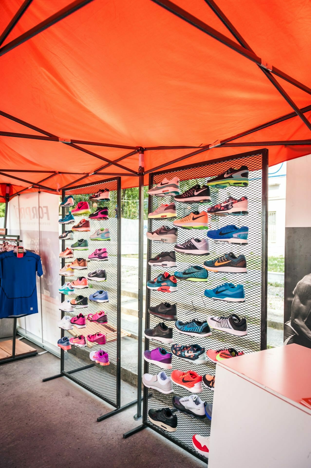 Photo report from the fair: An exhibition booth under an orange tent displays a wide range of colorful sports shoes. The booth is divided into several rows and columns, with shoes of different brands, designs and colors neatly arranged. On the left, a blue shirt is partially visible on the stand.  