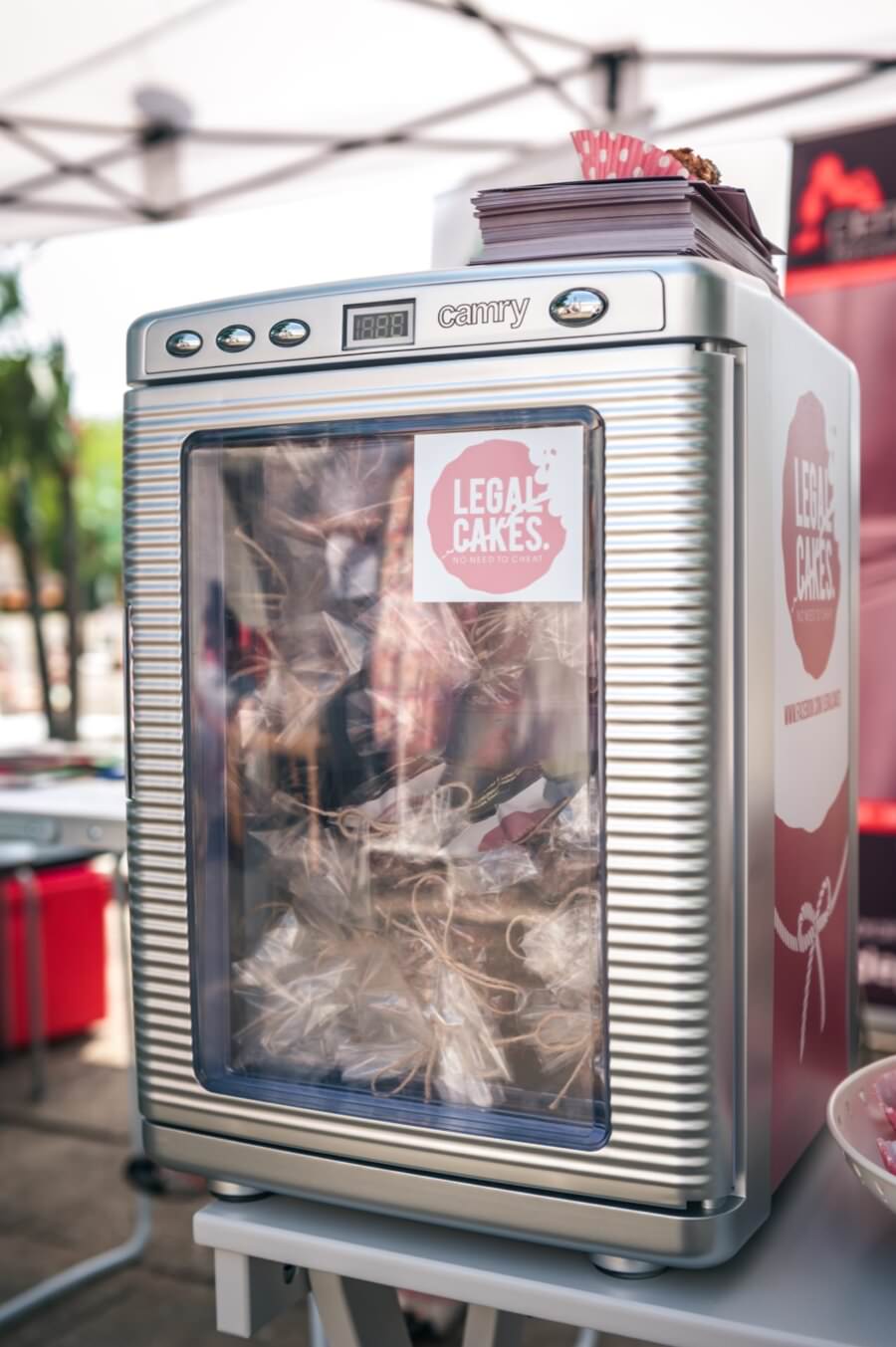 W srebrnej witrynie chłodniczej z napisem „Camry” znajdują się pakowane ciasta. Na drzwiach widnieje różowo-biały napis „Legalne ciasta”. Etui stanowi część fotorelacji z targów, ustawionej na stole w plenerze z rozmytym tłem.